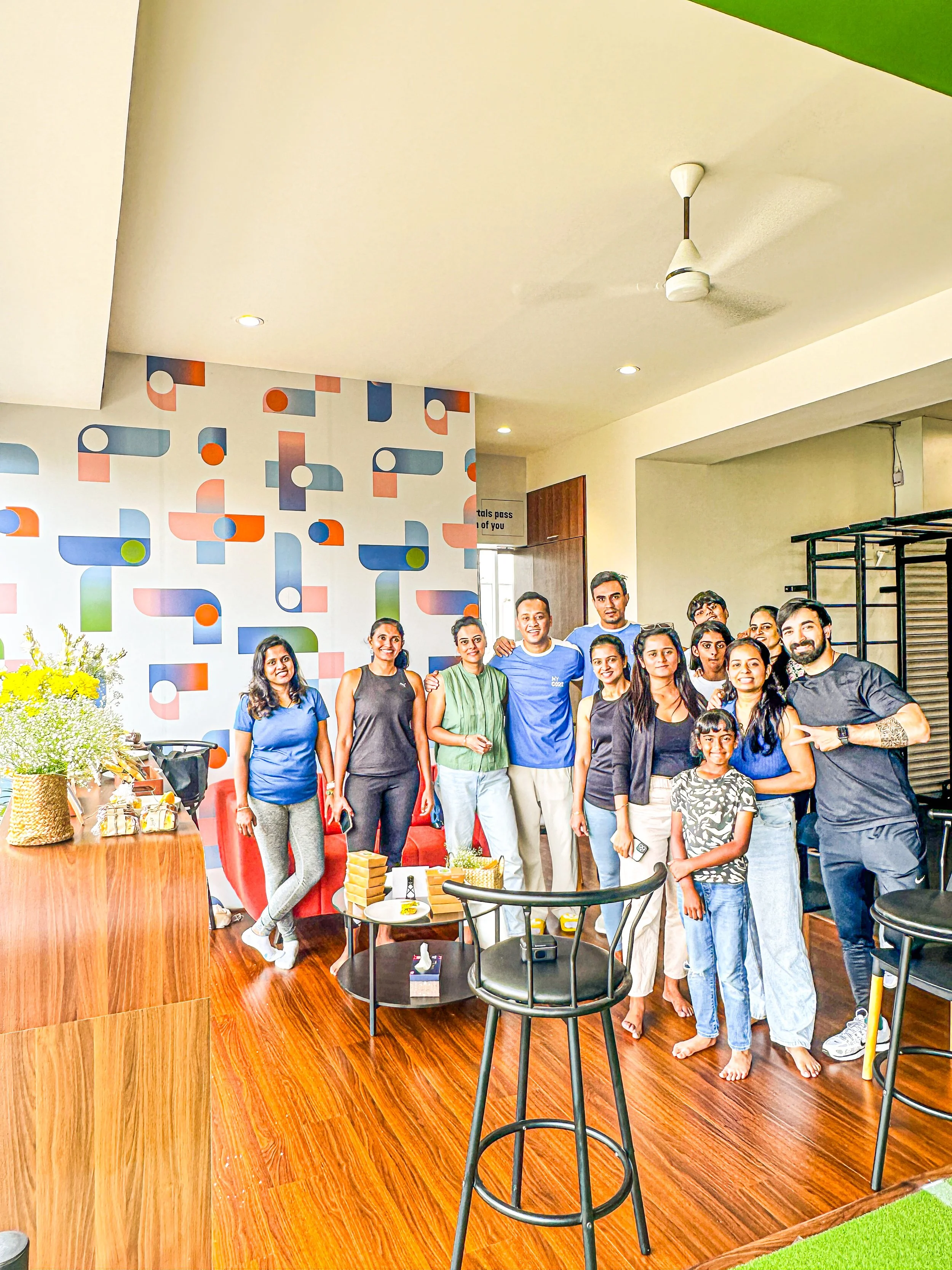 Group of people gathered in a modern indoor space, MyCore, for a celebration or gathering, with a colorful geometric wallpaper and wooden flooring.Train in a calm, small-batch strength and conditioning space in North Bengaluru, with personalised prog