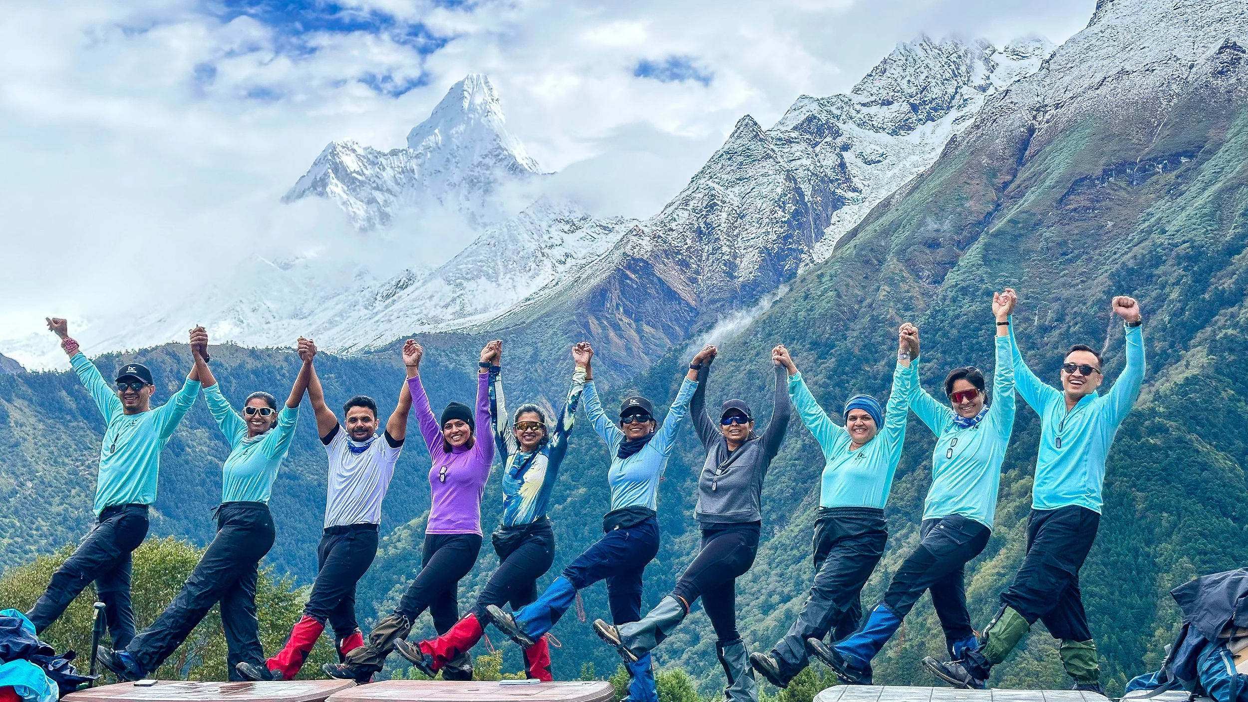 Group of nine people in outdoor gear holding hands and raising arms in front of a mountain landscape with snow-capped peaks, lush green mountains, and cloudy sky.