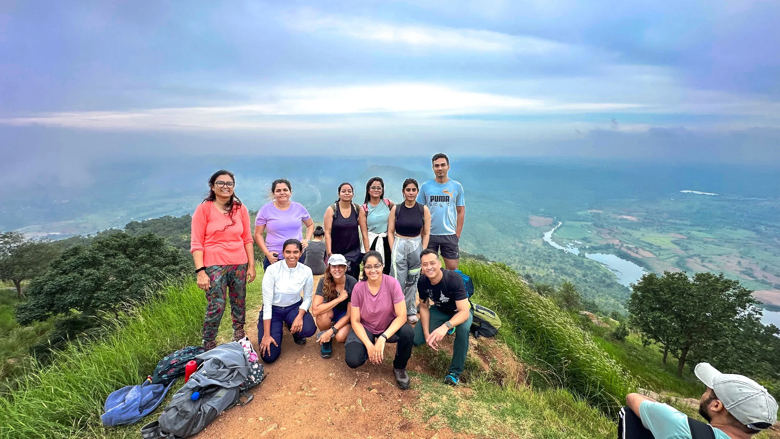 Group of hikers on a hill trail, near Bengaluru, overlooking a river valley under cloudy sky.