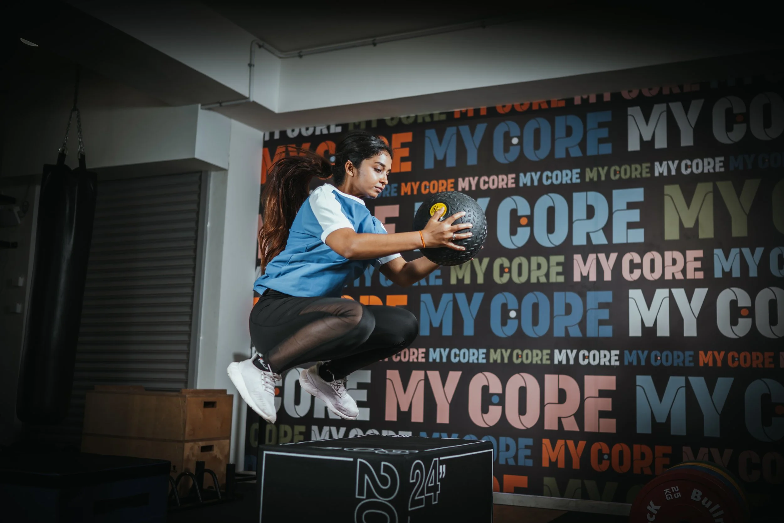 A woman in a blue and white athletic shirt and black leggings is jumping onto a plyometric box while holding a medicine ball in a gym with a colorful "MY CORE" themed wall in the background.