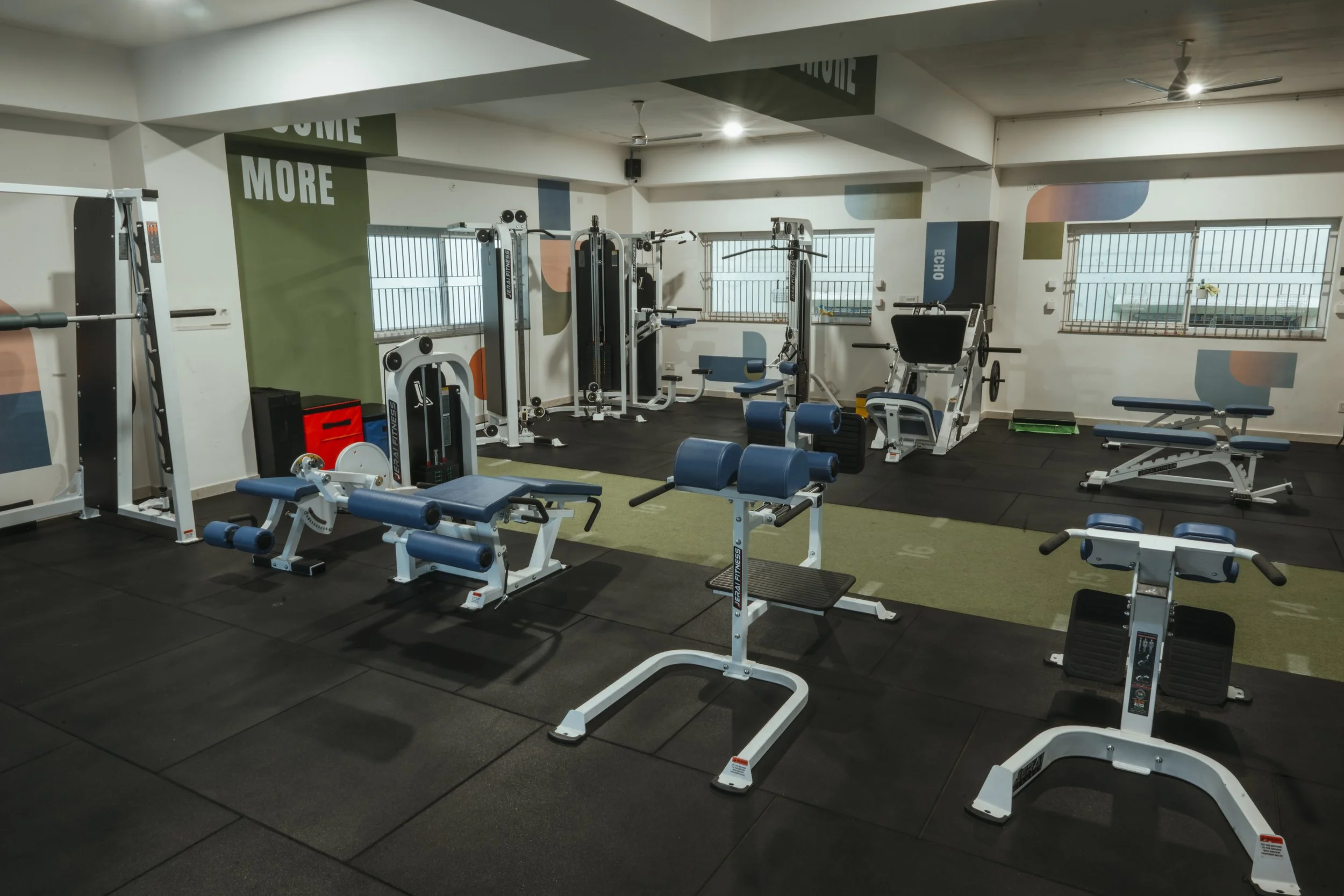 Empty gym with various workout equipment including weight benches, resistance machines, and cardio equipment, with windows letting in natural light. The space is MyCore, a gym in North Benghaluru 