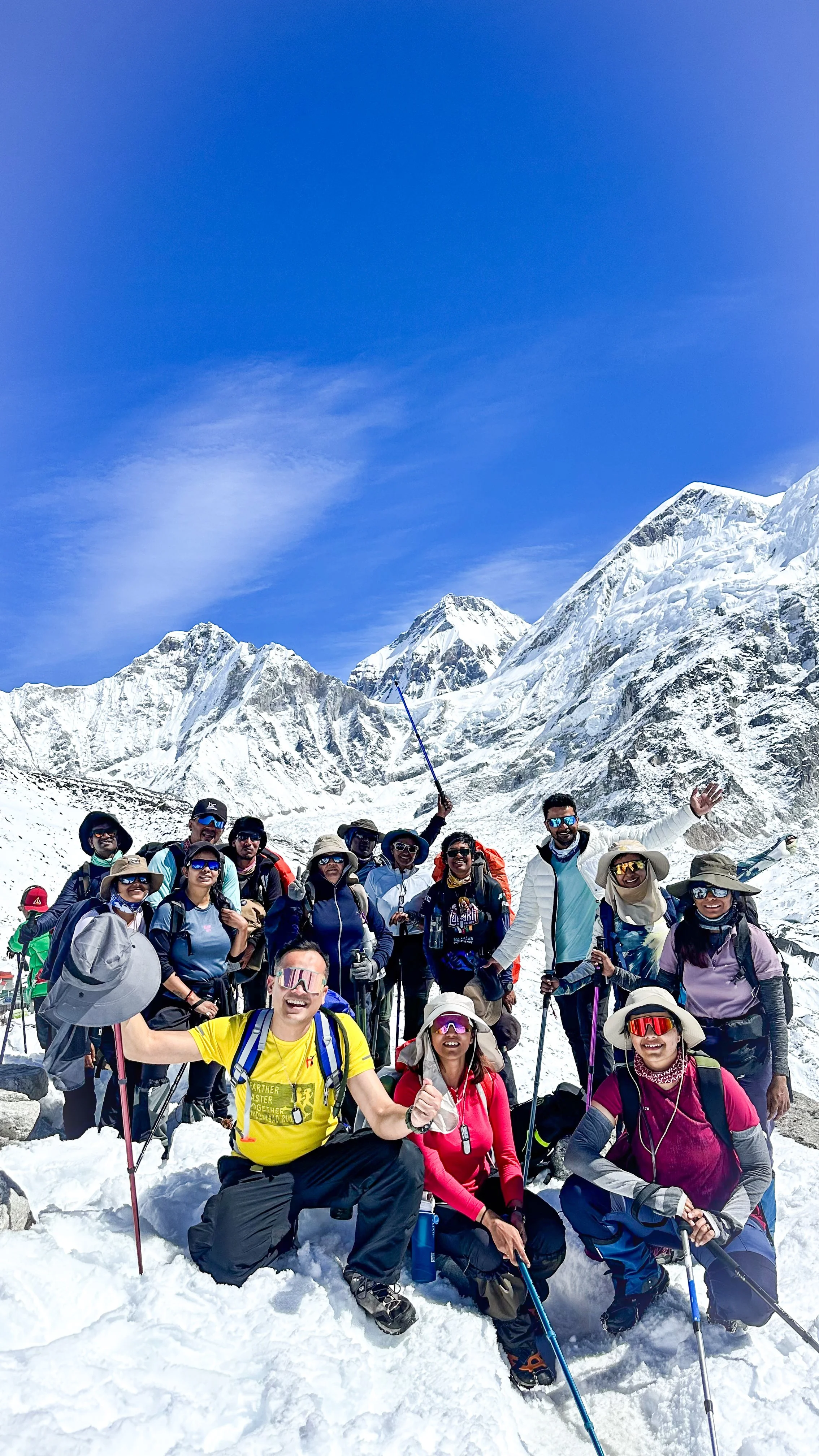Group of mountaineers in snowy mountain landscape, some smiling and holding hiking poles, with snow-covered peaks and clear blue sky in background.
