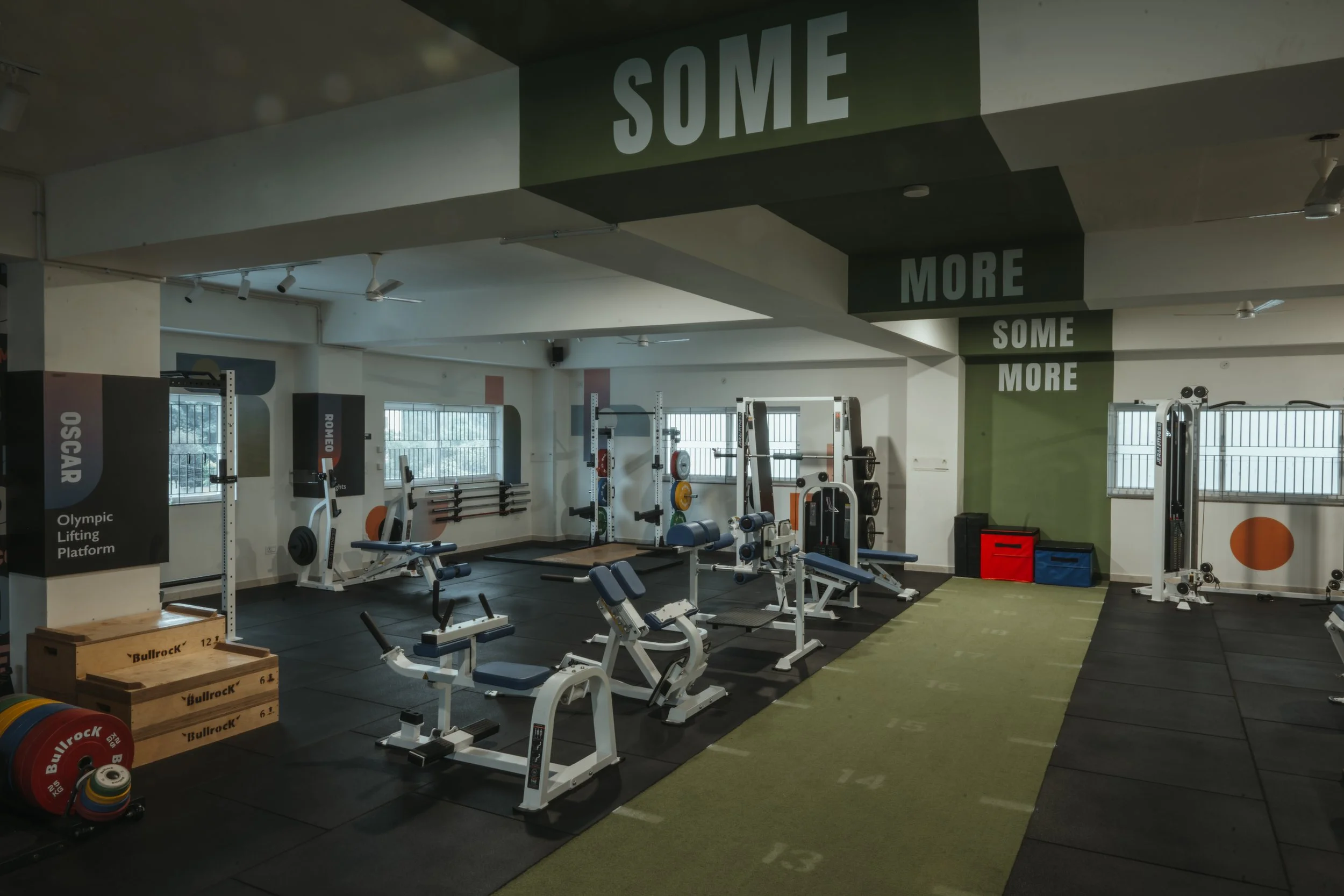 Empty gym with various exercise equipment including weightlifting machines, benches, and weights, featuring motivational words 'SOME MORE' on the ceiling and walls. The space is MyCore, a gym in North Benghaluru 