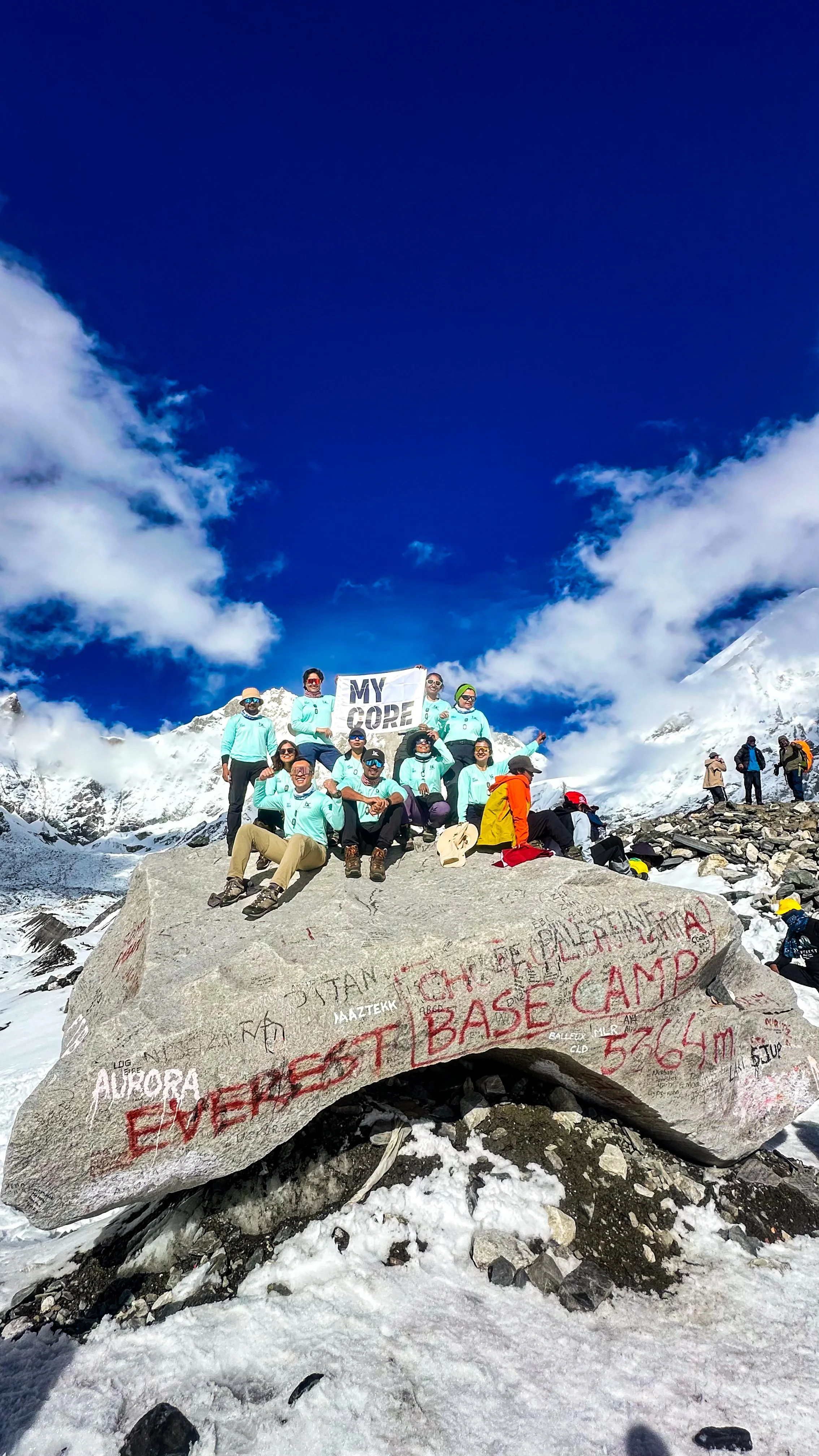 Group of climbers at Everest Base Camp on a snowy mountain with clouds and blue sky, holding a sign that says 'MY CORE'.