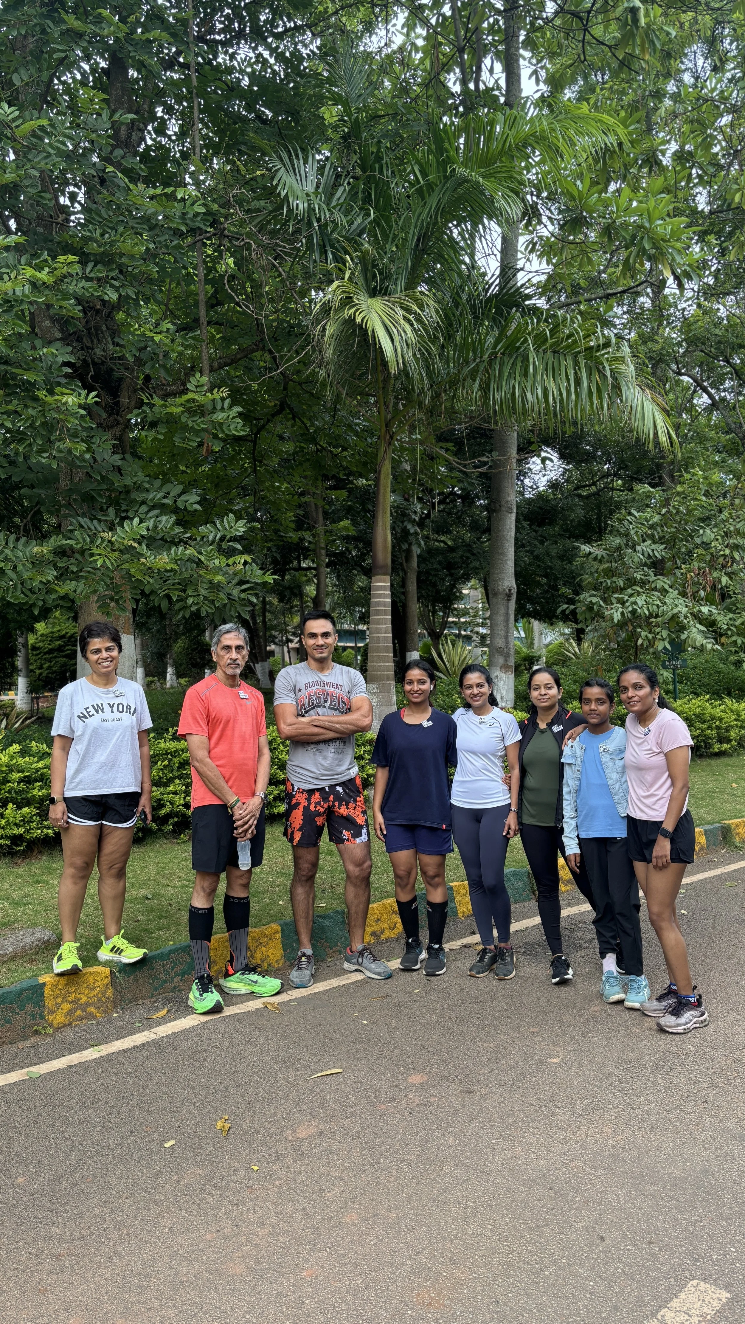 Group of eight people standing outdoors on a paved path, surrounded by green trees and plants, posing for a photo.