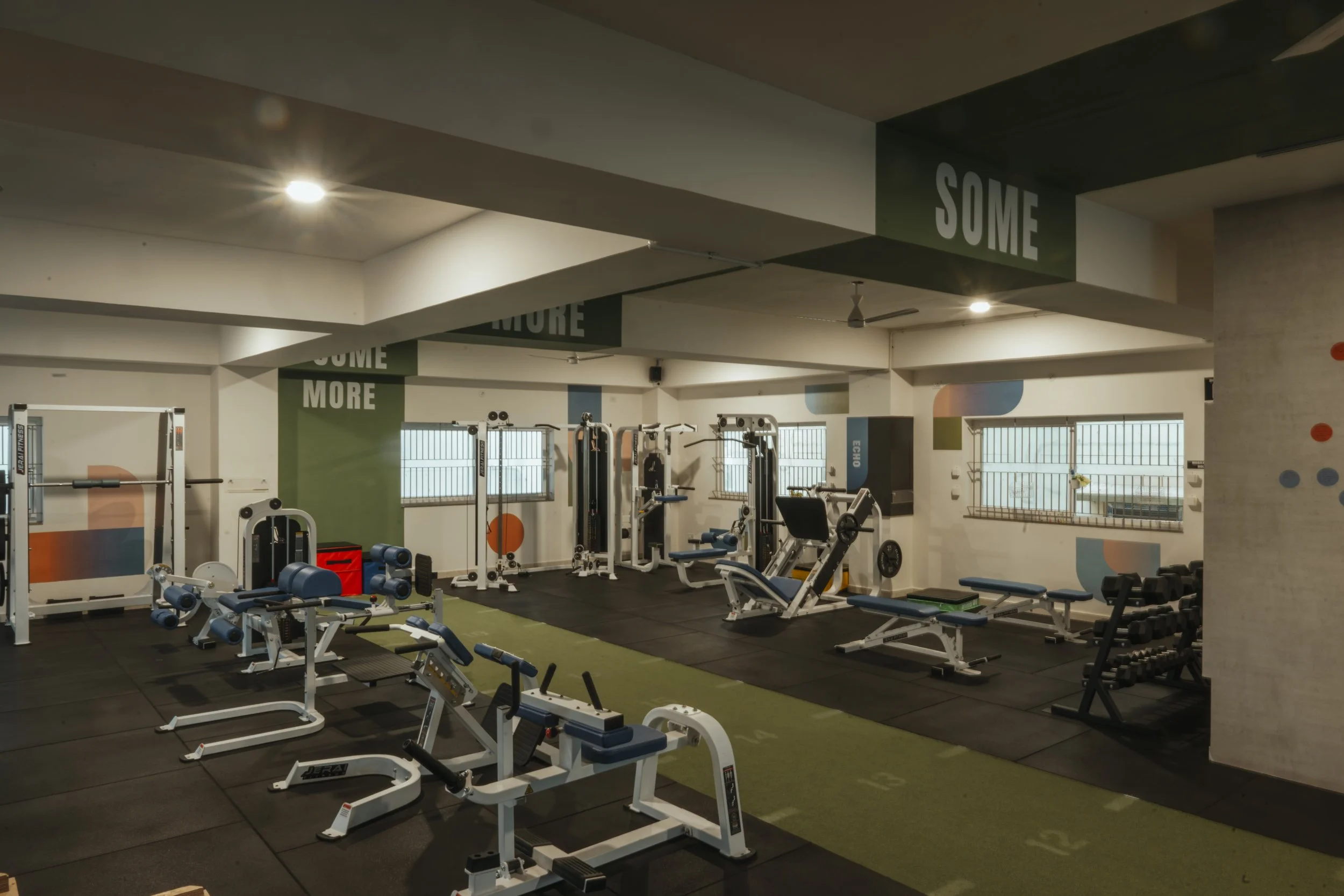 Empty gym with various fitness equipment including weights, benches, and machines, with windows and motivational signs that read 'SOME MORE'.