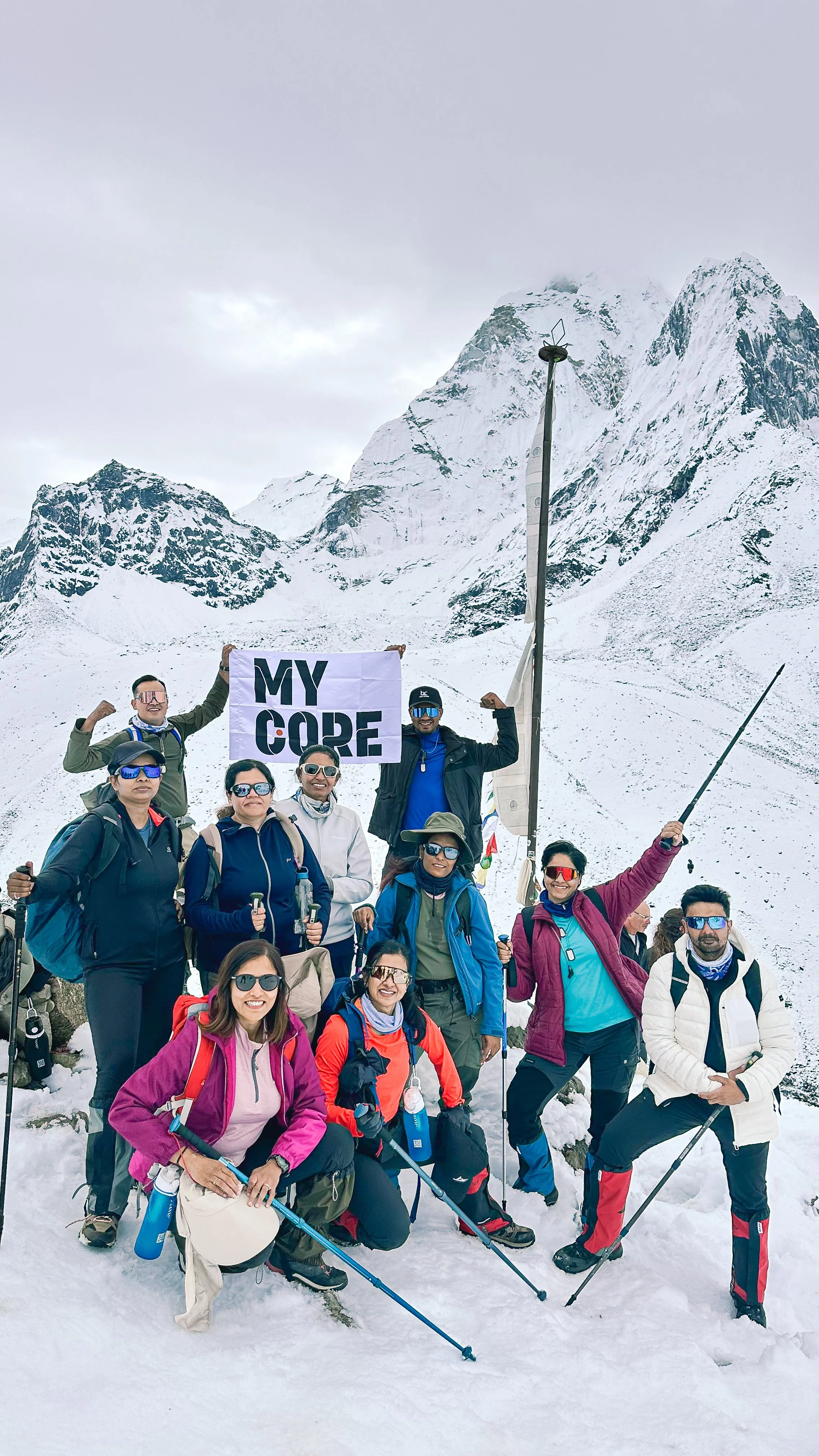 Group of ten hikers in winter gear on snowy mountain with snowy peaks in the background. Some are smiling, posing, and holding trekking poles, with one person holding a sign that reads 'MY CORE' and another holding a flag.