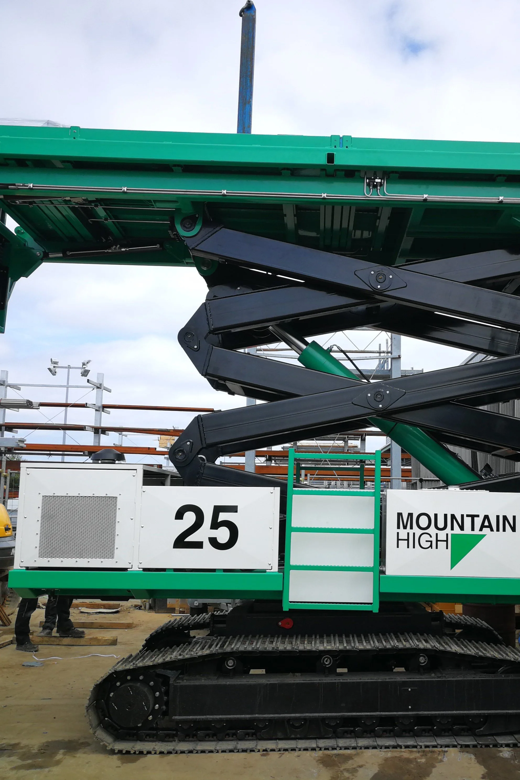 Een bouwmachine met een groen en zwart chassis die zich op een bouwplaats bevindt, met een bouwbord waarop '25' en 'MOUNTAIN HIGH' staat.