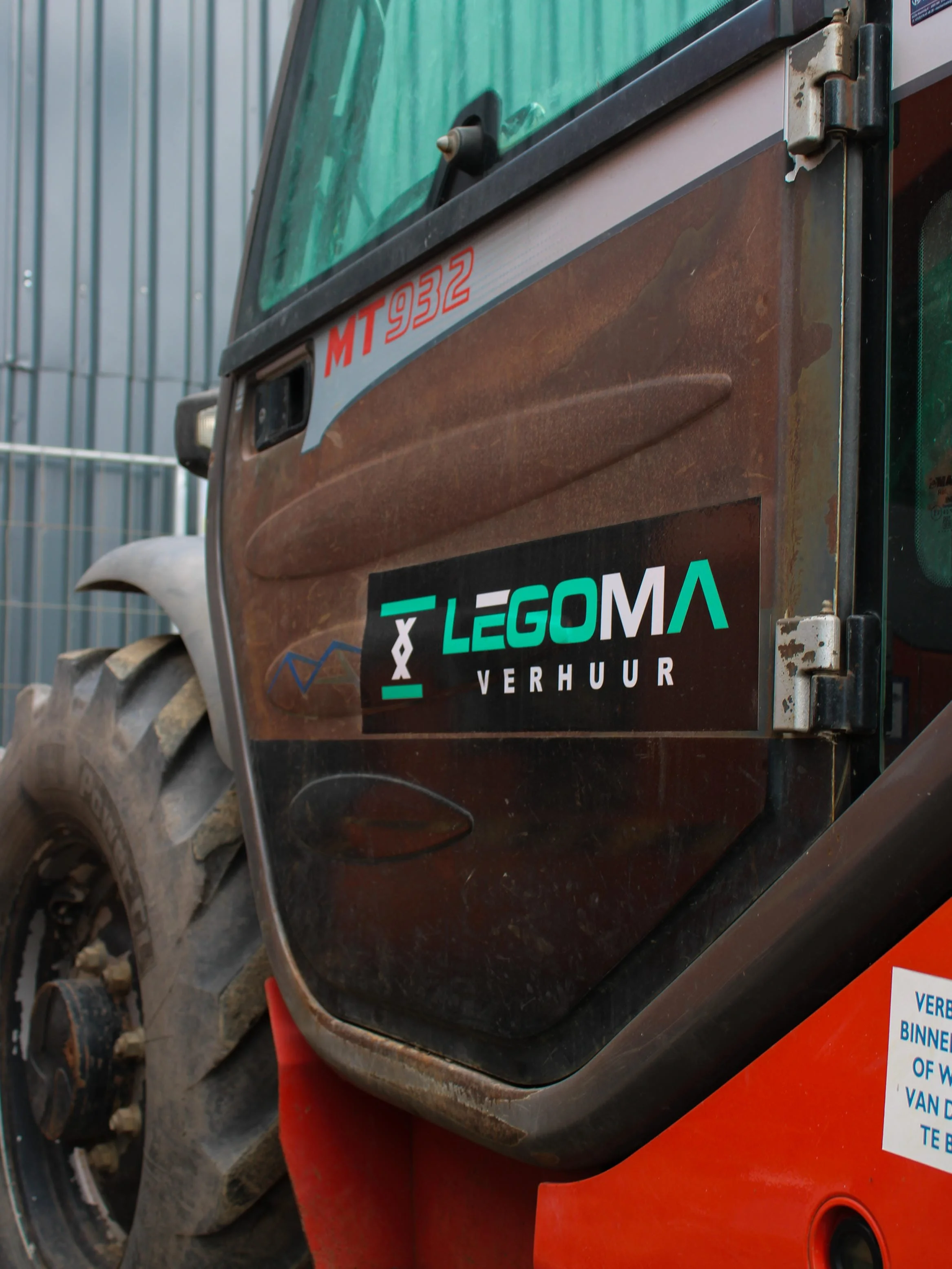 Close-up van de voorkant van een oude bulldozer met een sticker van ELGOMA Verhuur. De hijskraan is roestig en vuil, met grote banden op de achtergrond.