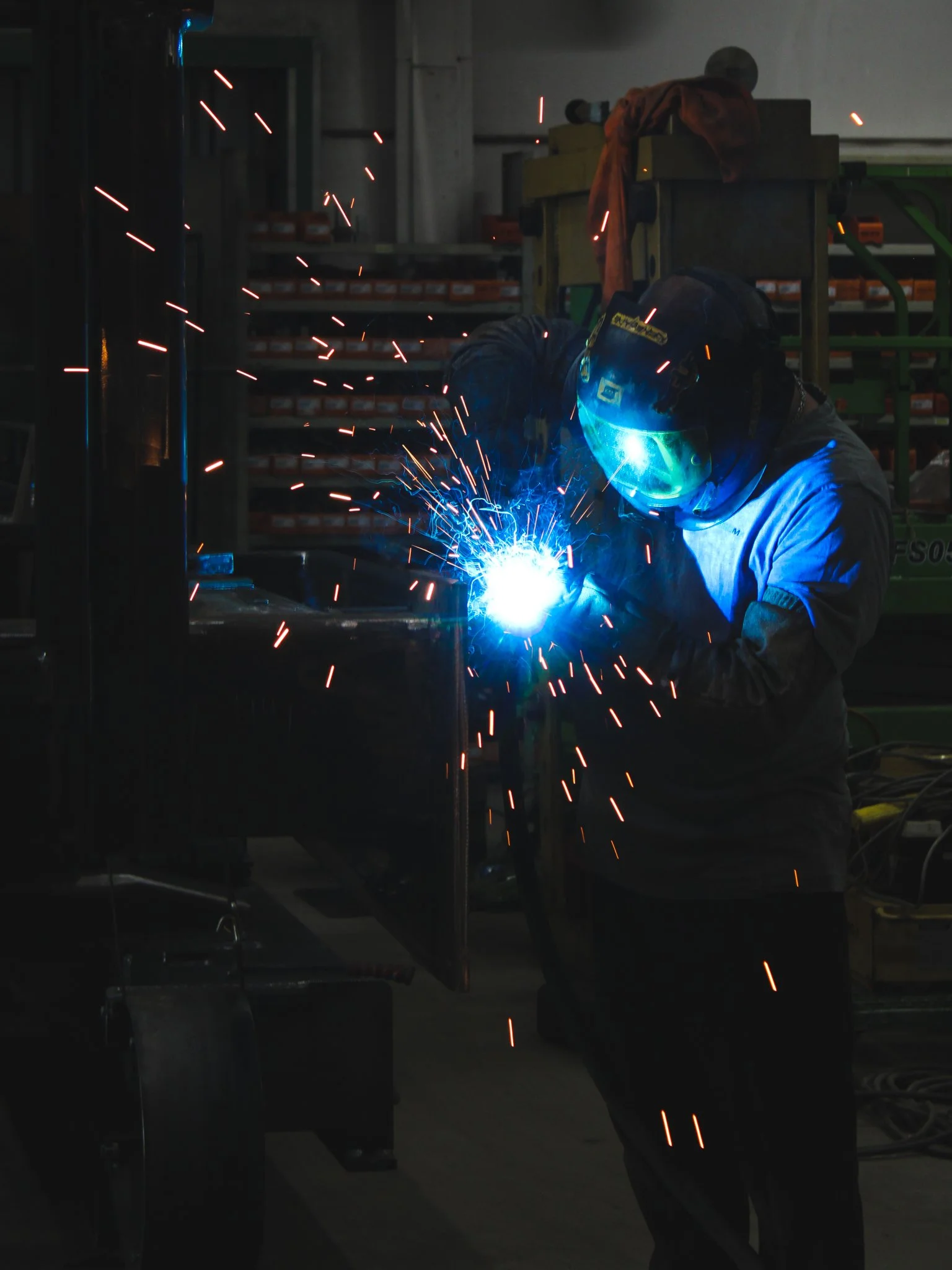 Een werknemer die laswerk uitvoert, met een beschermende helm en een lasbril, in een werkplaats met metalen rekken en gereedschap.