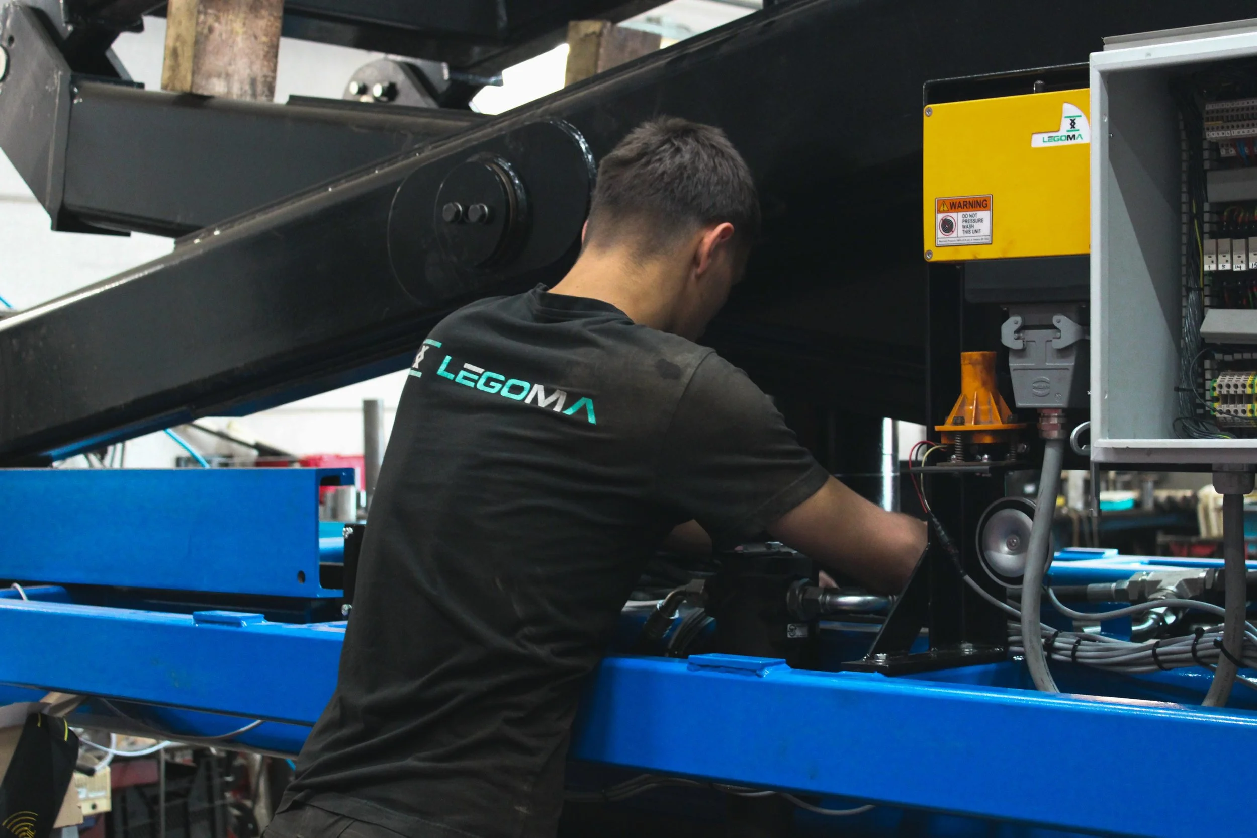 Man werkt aan technische apparatuur in een werkplaats, draagt een zwart shirt met het logo 'LEGOMA' op de achterkant.