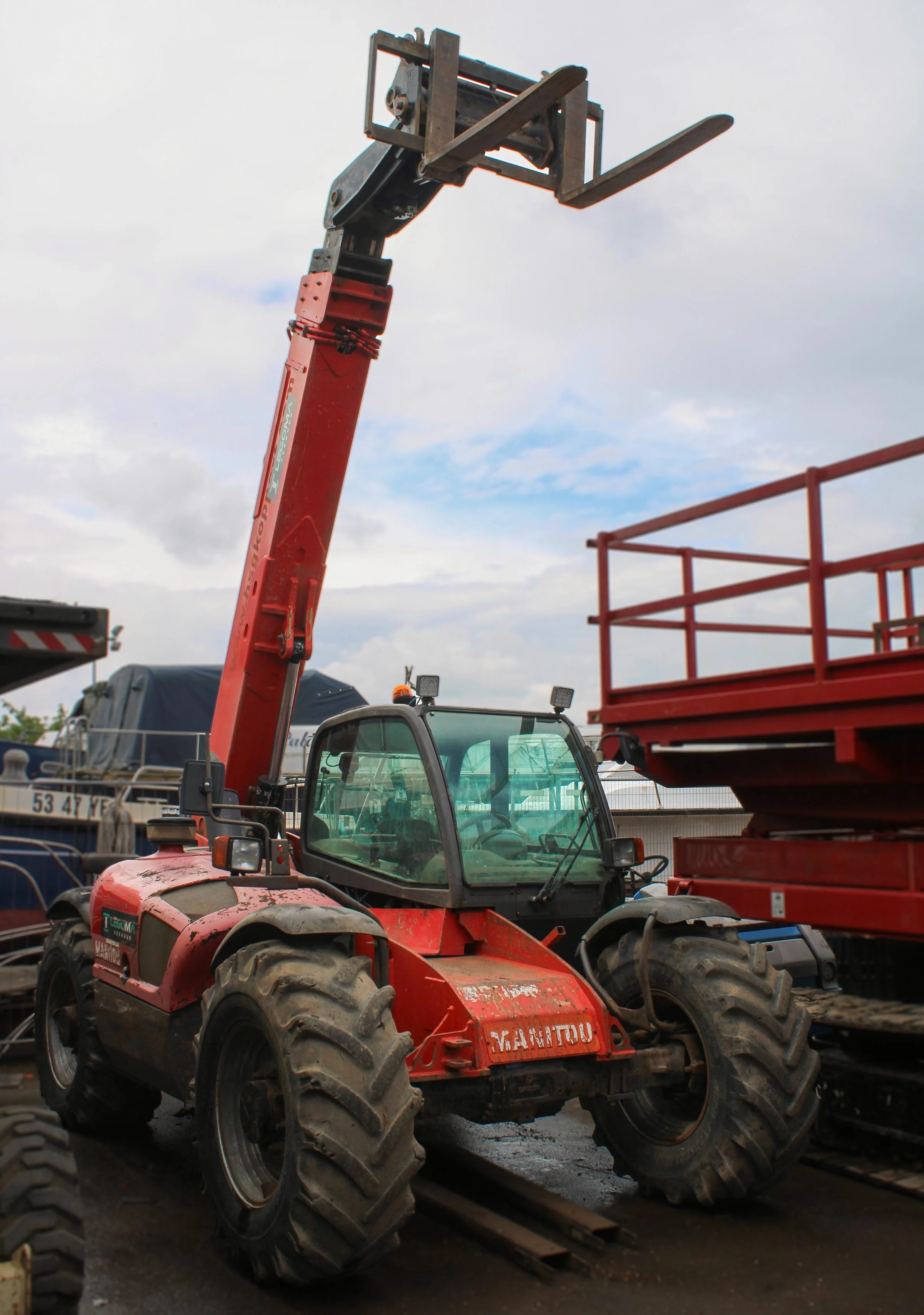 Een rode Manitou heftractor met een uitgestrekte hefarm, geparkeerd op een betonnen ondergrond met hoogwerker erachter