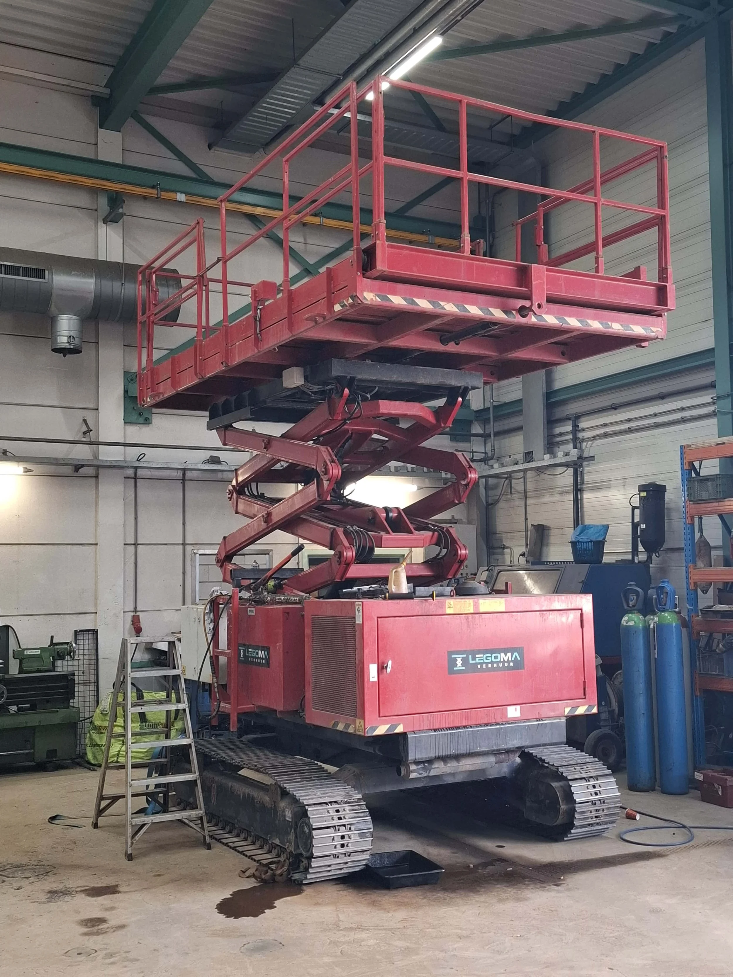 Een rediombouwmachine in een werkplaats. De machine is rood en heeft een uitschuifbare platform met een hefstructuur. Er ligt een klein plas water op de vloer onder de machine, en er staat een ladder naast.