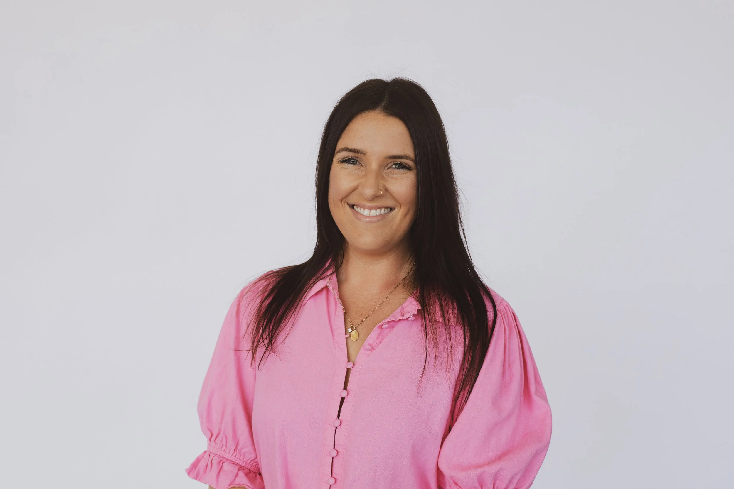 Portrait of a woman with long dark hair wearing a pink blouse and smiling against a plain background.