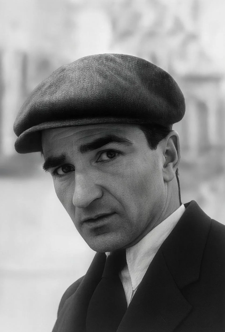 Siyah-beyaz fotoğraf, bir erkeğin yüzü ve omuzlarını gösteriyor. Erkek, koyu renkli sarkık bir beret giymiş ve ciddi bir ifadesi var. Arka planda belirsiz yapılar bulunuyor.