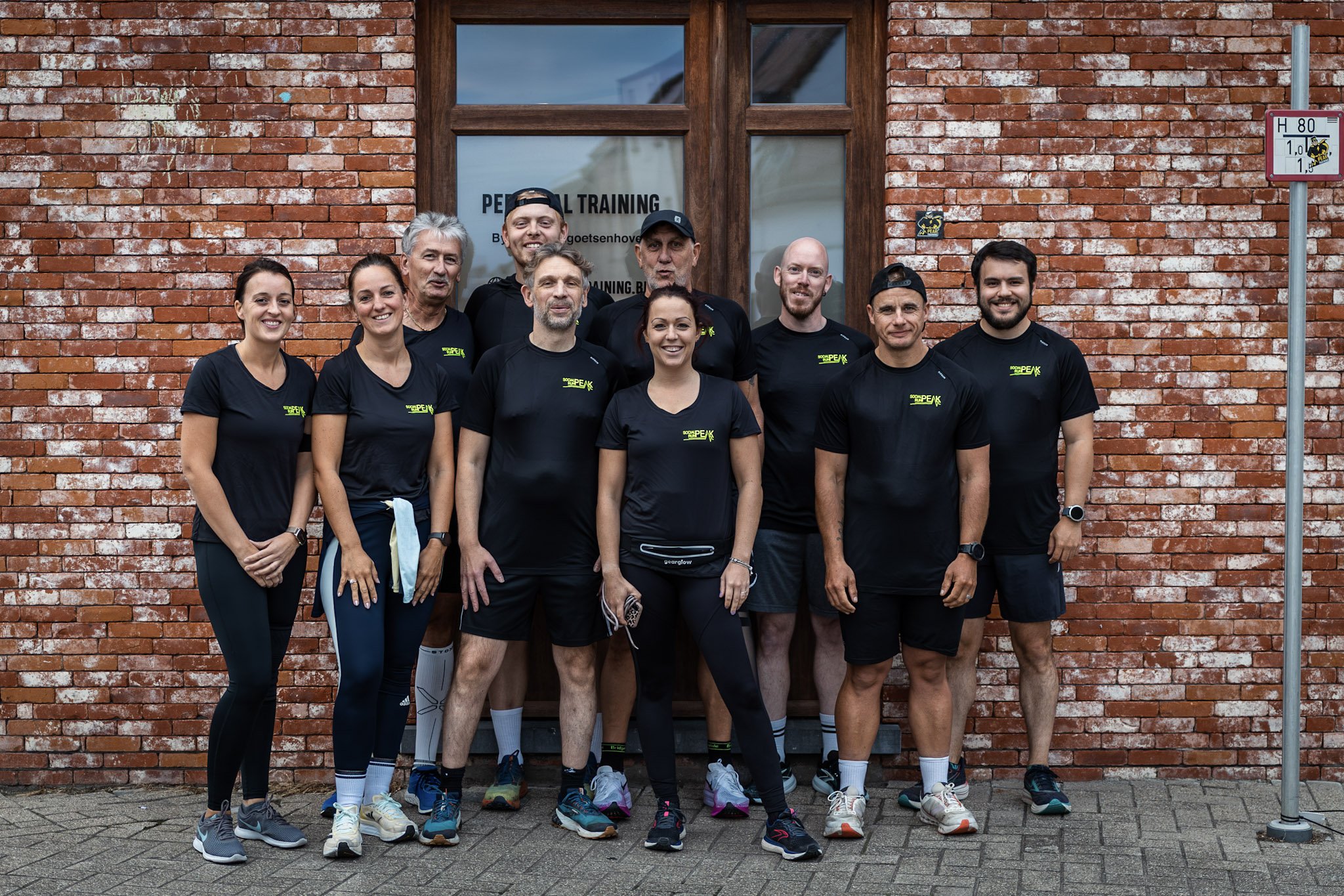 Een groep van tien lopers in zwart sportkleding poseert voor een bakstenen muur met een houten deur. Ze lijken zich voor te bereiden op of net klaar te zijn met een trainingssessie.