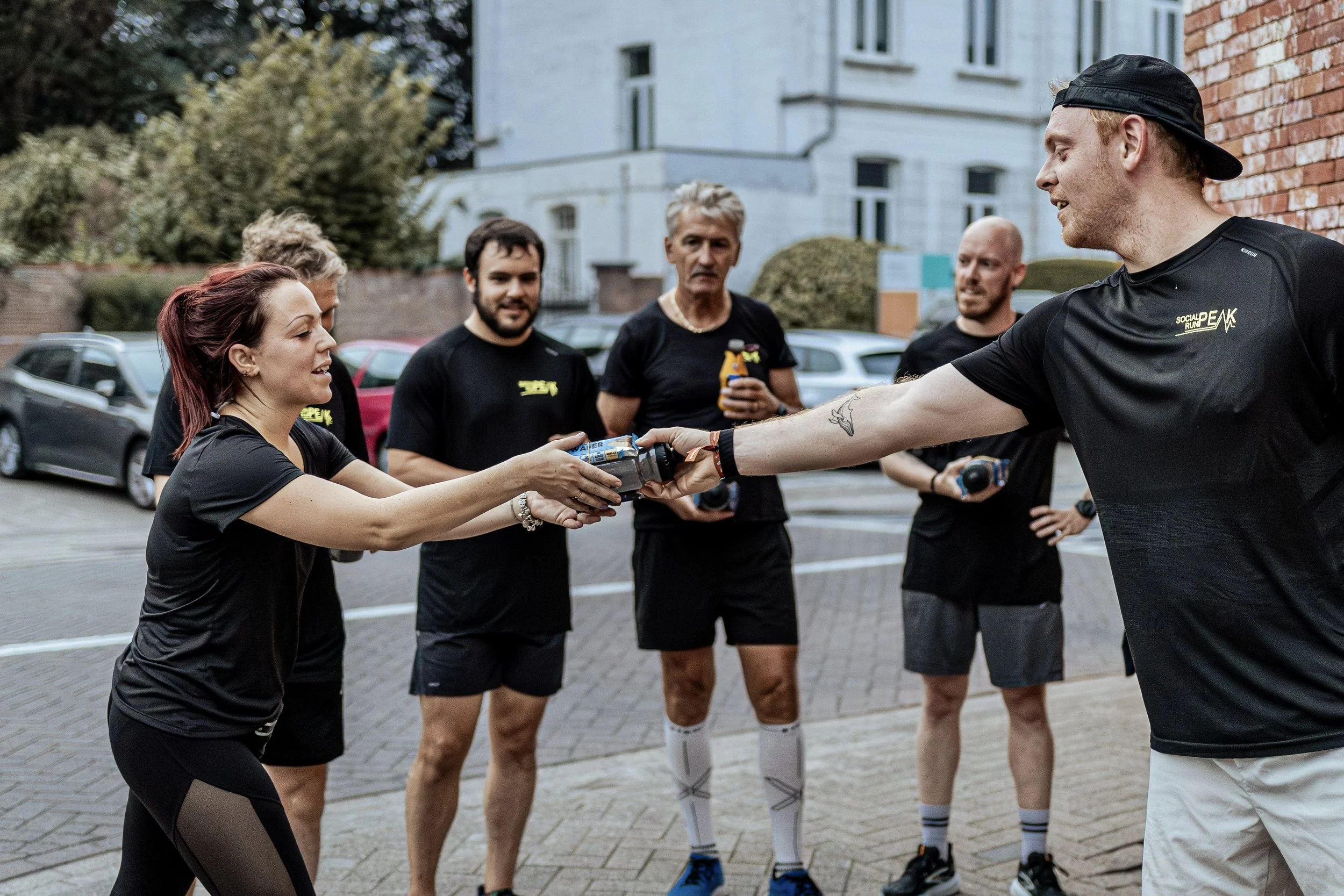Vrouw krijgt een waterflesje van een man tijdens een outdoor loopgroep bij rustige straat met auto's en huizen op achtergrond.