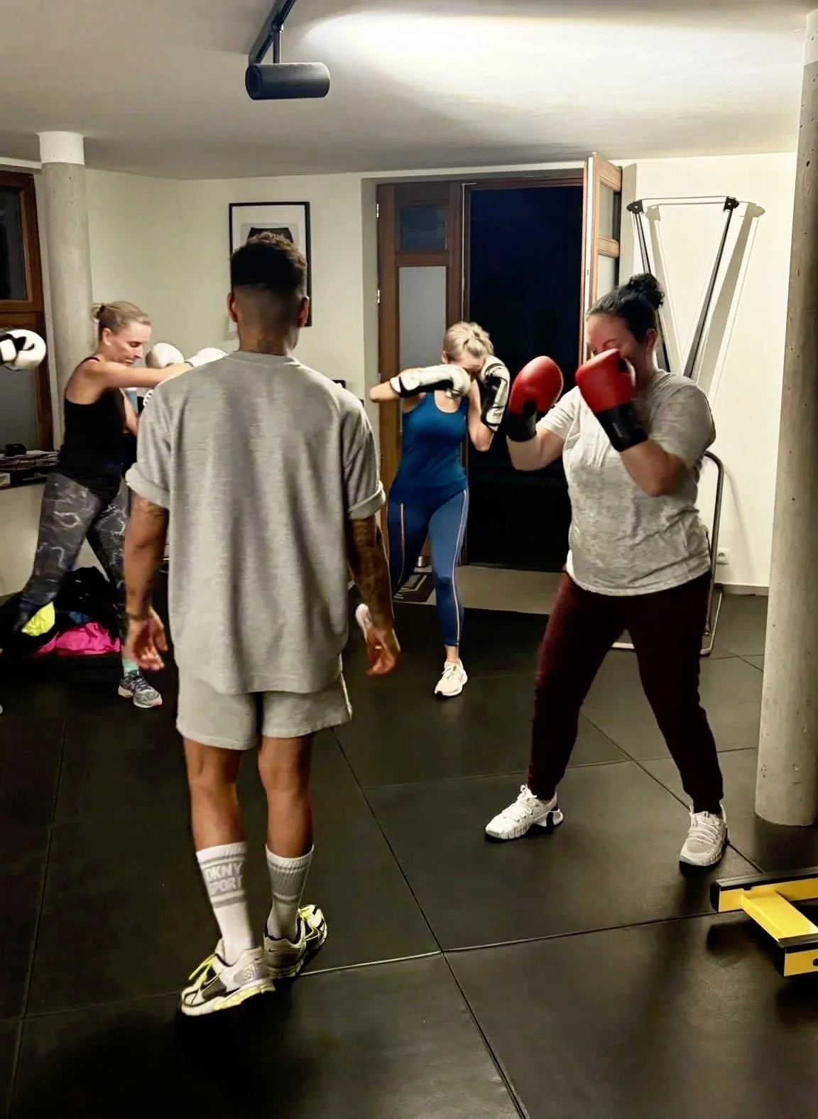 Four people practicing boxing in a gym, with one woman wearing red gloves and gloves, and others in athletic clothing, inside a room with black flooring and workout equipment.