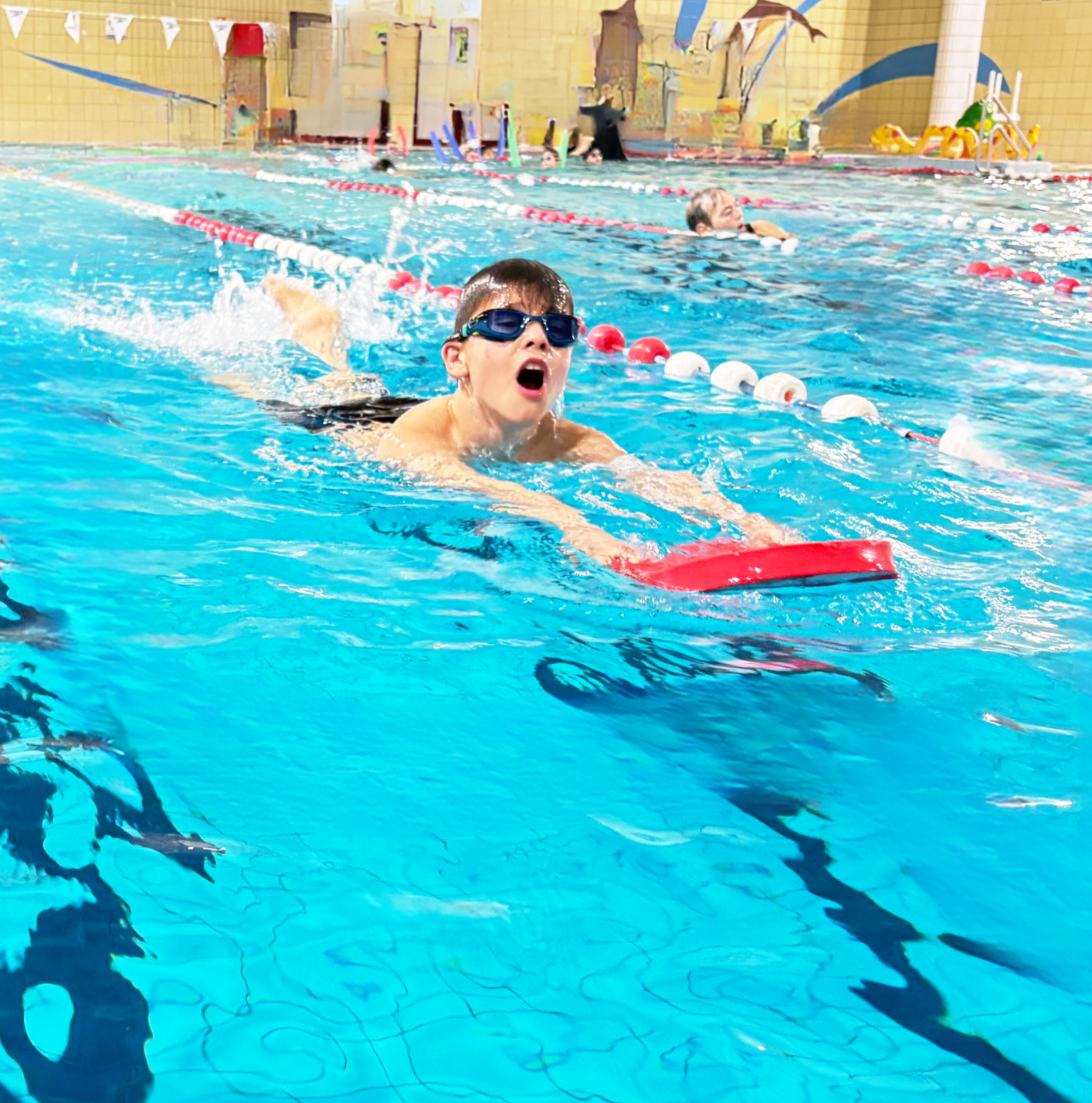 A young boy with goggles swimming in an indoor swimming pool using a kickboard, with other swimmers and aquatic equipment visible in the background.