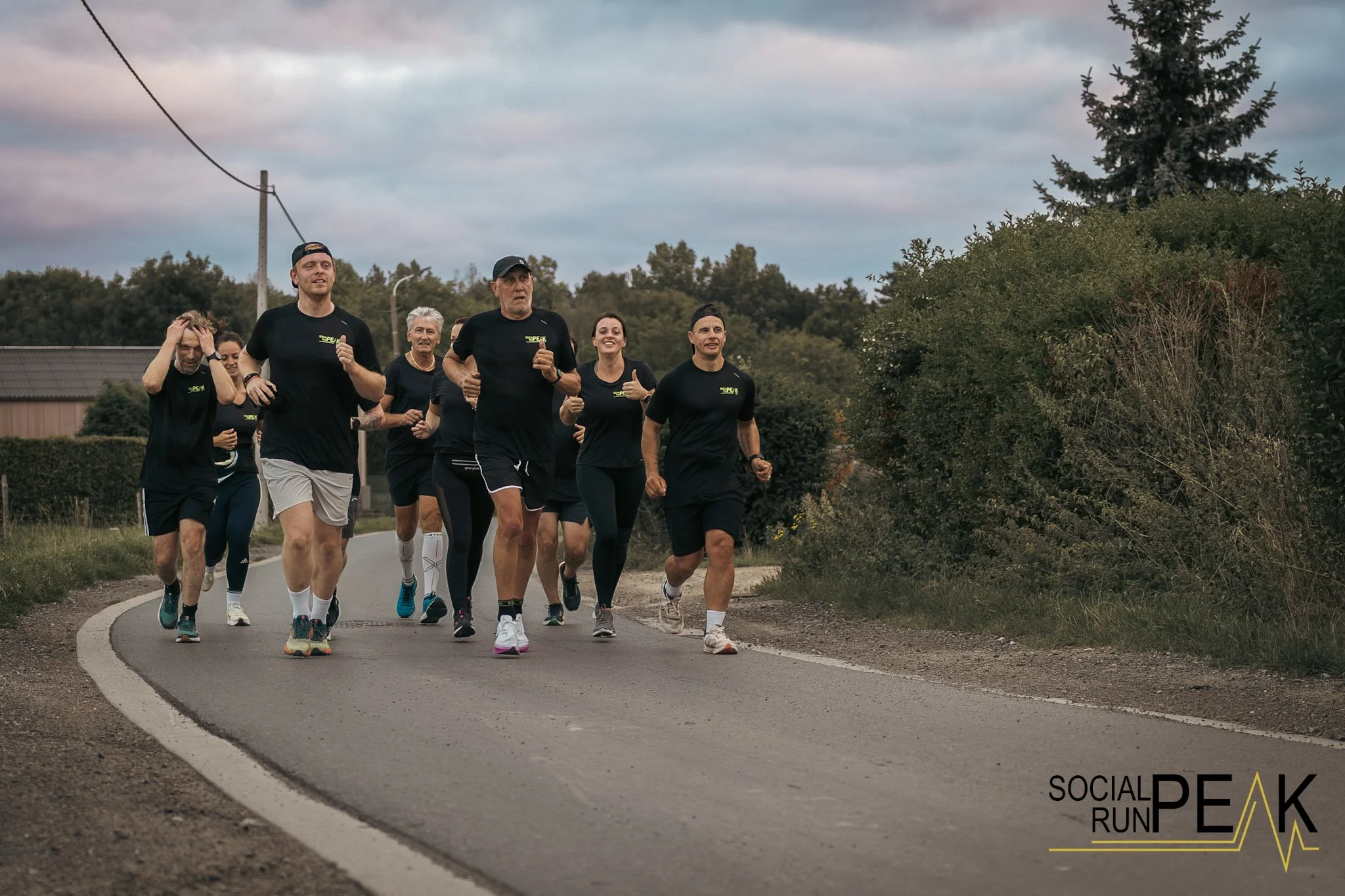 Een groep mensen die meedoet aan een hardloopevenement op een landelijke weg, gekleed in zwarte sportkleding met 'Social Run Peak' branding, onder een bewolkte hemel.