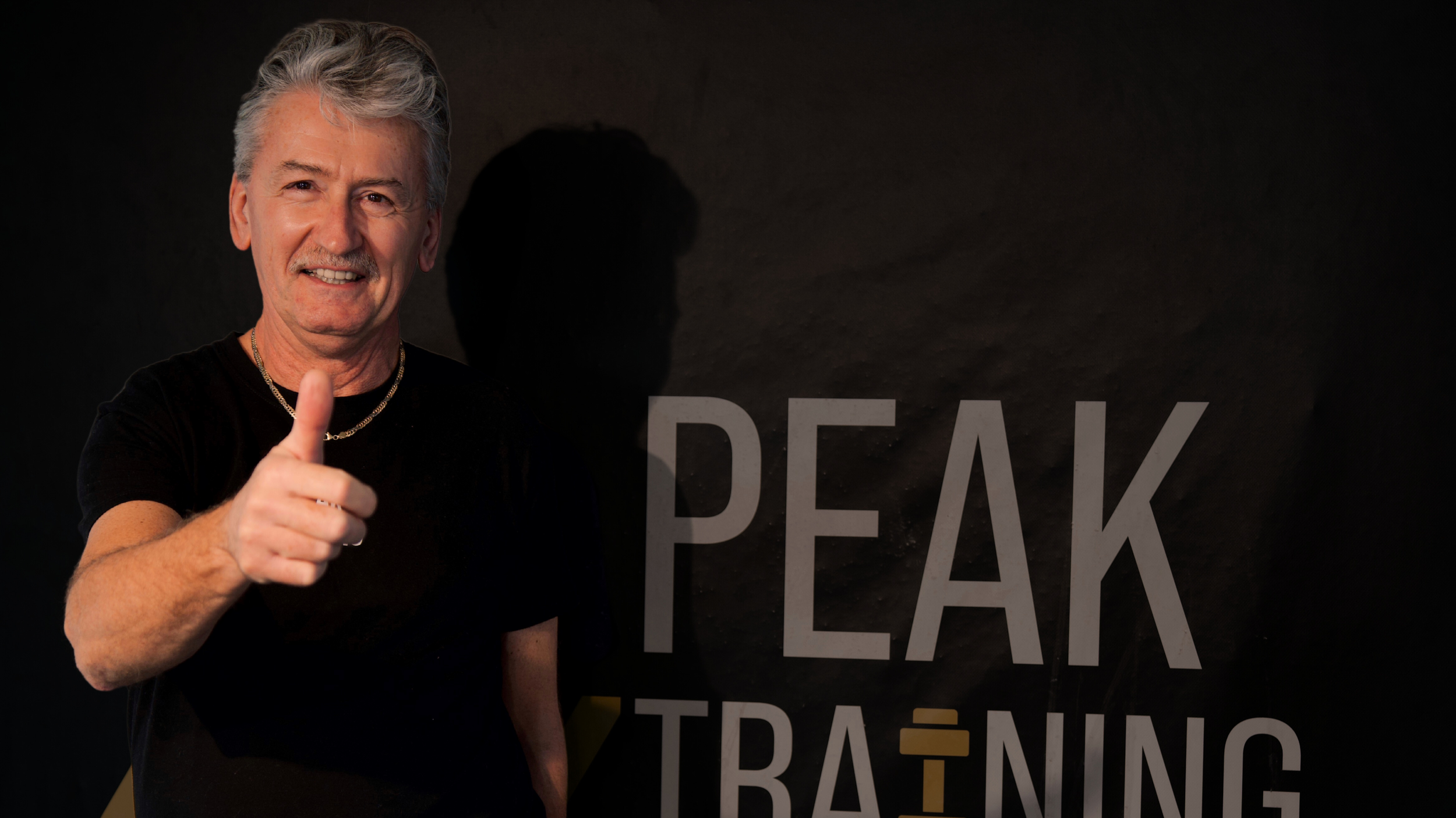Man met grijze haar geeft duim omhoog voor een zwarte achtergrond met de tekst 'PEAK TRAINING'.