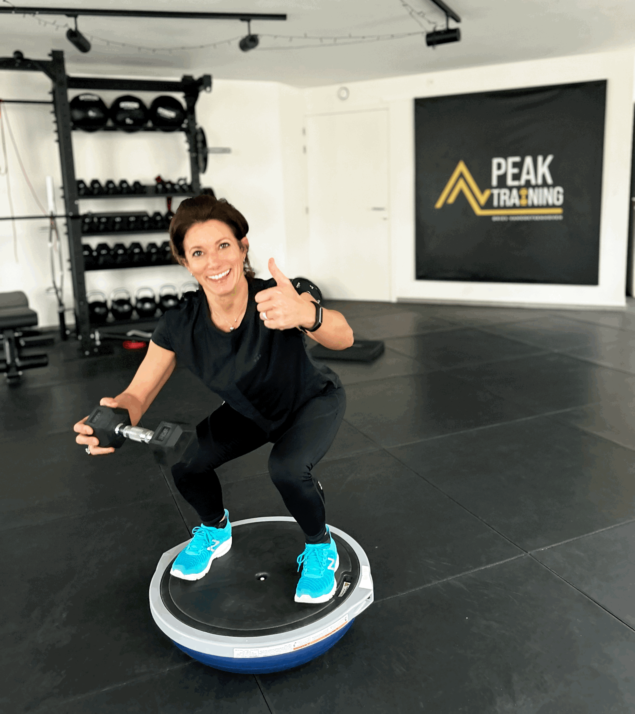 A woman smiling and giving a thumbs up while holding a dumbbell, standing on a balance trainer in a gym with weights on shelves and a sign that reads 'Peak Training' in the background.