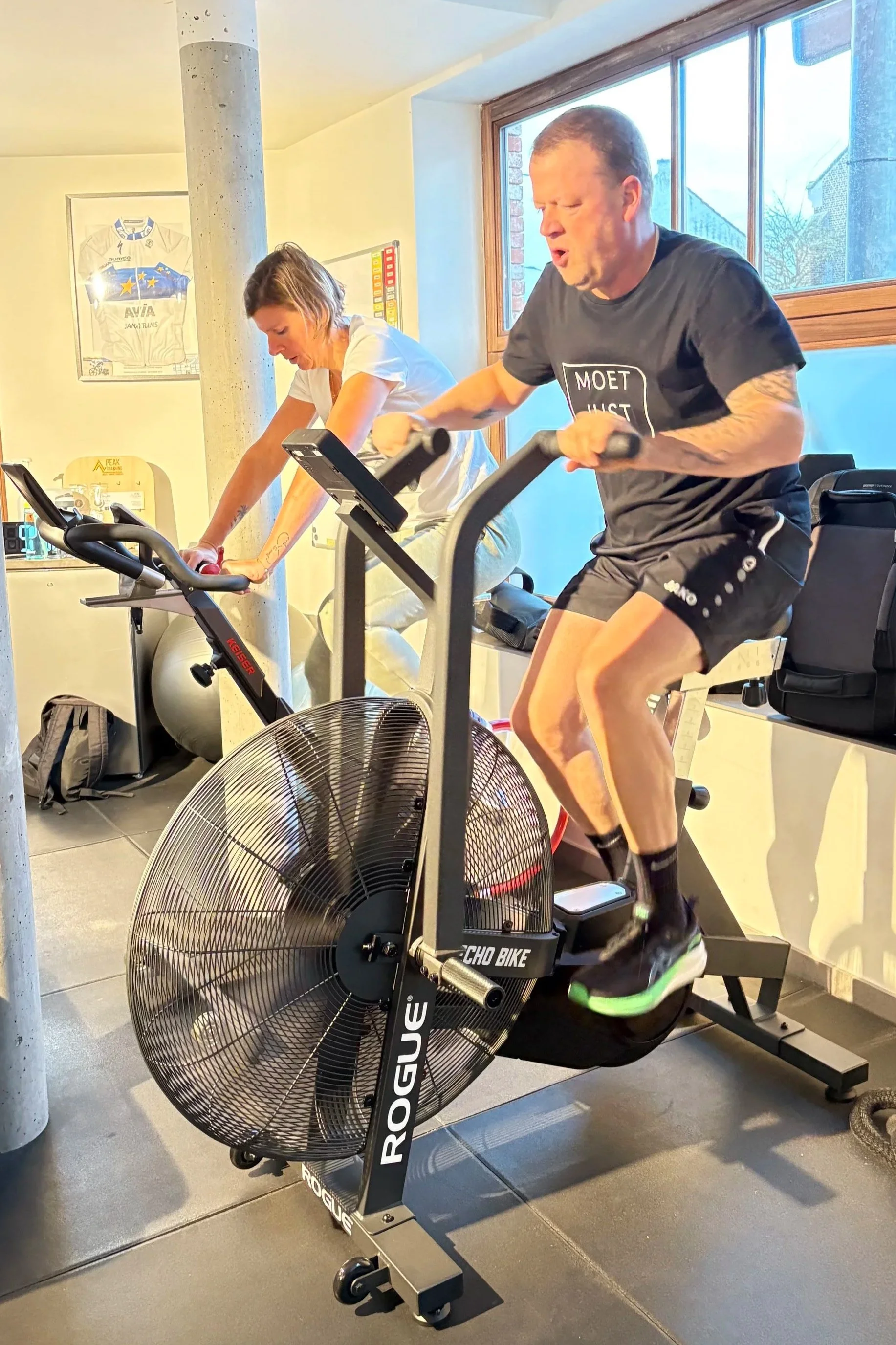 Two people exercising indoors on stationary bikes, with a woman on the bike in the background and a man in the foreground, with a large window behind them showing a building outside.