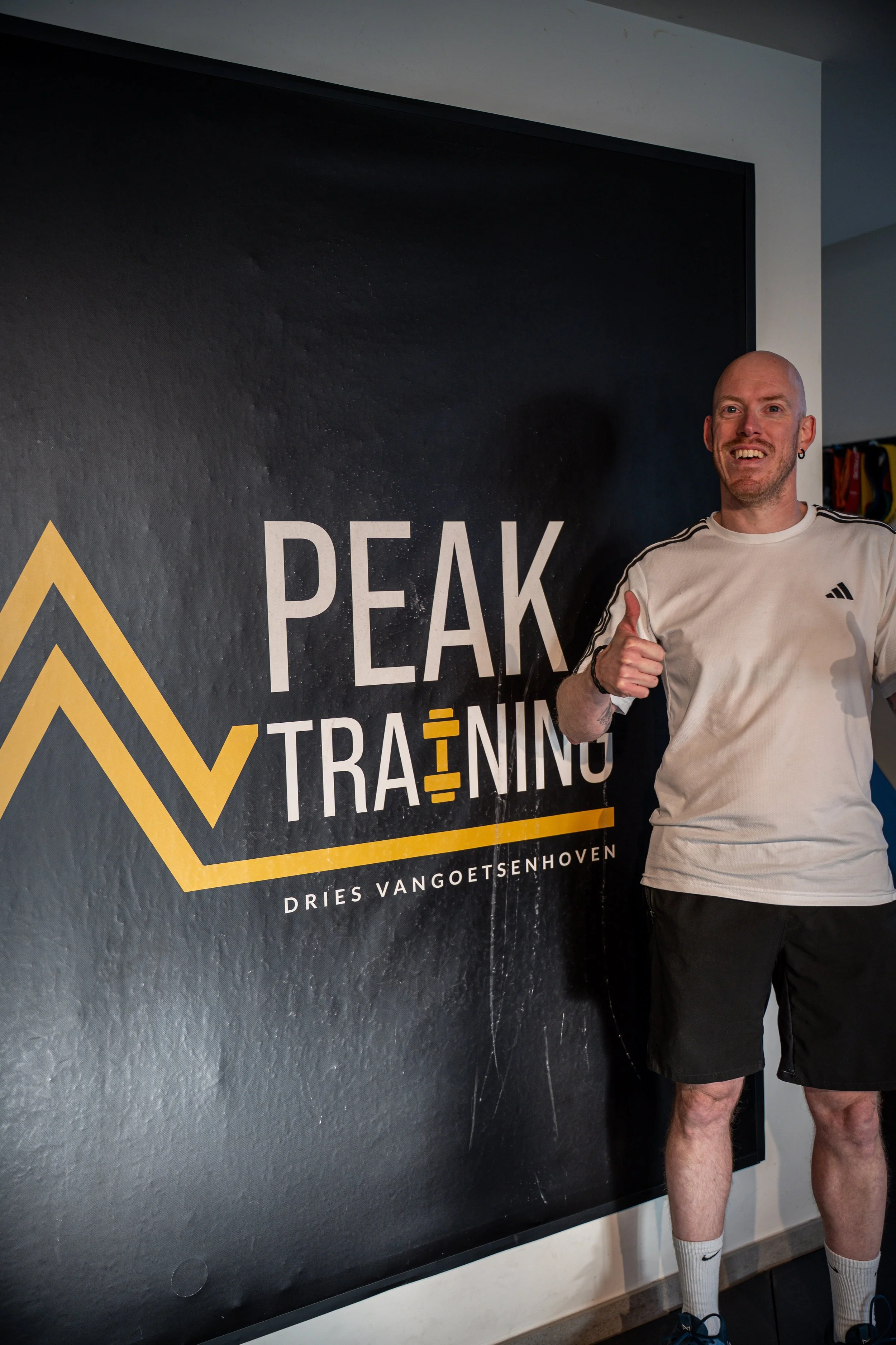 Man in sportkleding staat naast een PEAK TRAINING bord, geeft een duim omhoog en glimlacht.