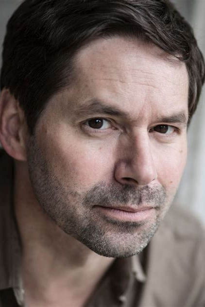 Dietmar Horcicka - Dozent der internationalen Schauspiel-Akademie für Film und Theater in Hamburg. Nahaufnahme eines Mannes mit dunklem Haar, grauer Kleidung, schrägem Blick und leichtem Lächeln.