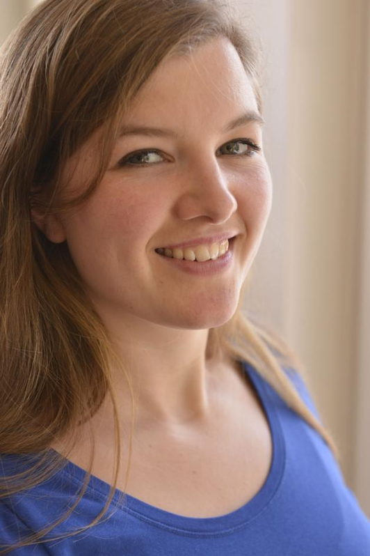 Lächelnde junge Frau mit braunen Haaren in einem blauen Oberteil vor einer hellen Wand.
