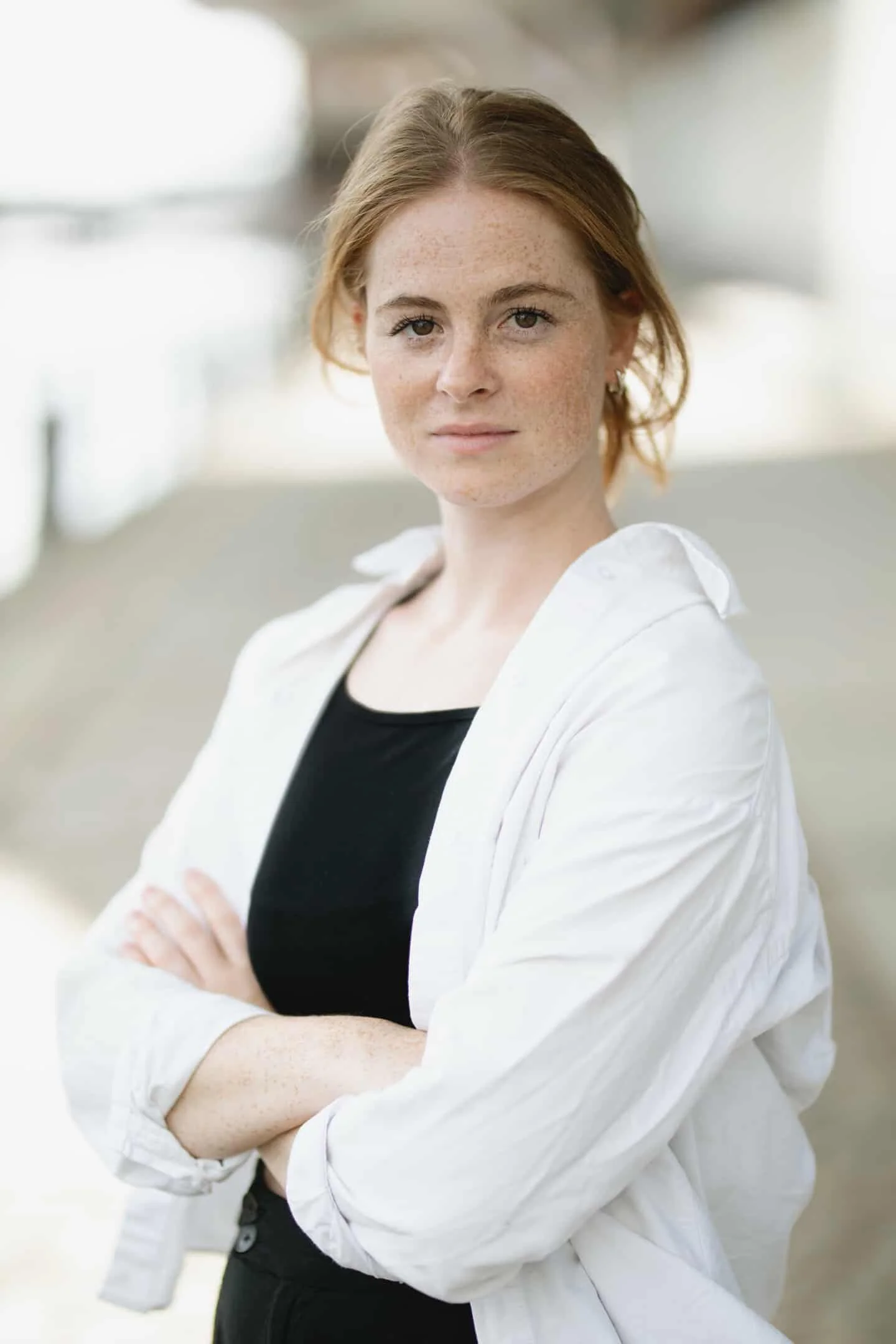Anke Bautzmann - ehemalige Absolventin internationalen Schauspiel-Akademie. Junge Frau mit roten Haaren und Sommersprossen, trägt eine weiße Jacke und ein schwarzes Oberteil, vor einem grauen und weißen Hintergrund.