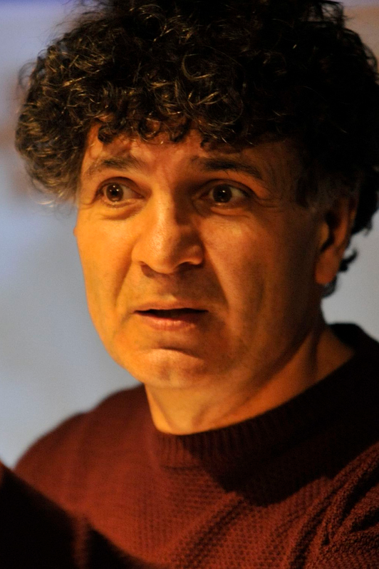 Mahmut Canbay - Dozent der internationalen Schauspiel-Akademie für Film und Theater in Hamburg. Nahaufnahme eines Mannes mit lockigem dunklem Haar, der einen schwarzen Pullover trägt, blickt nach links und hat einen nachdenklichen Ausdruck.