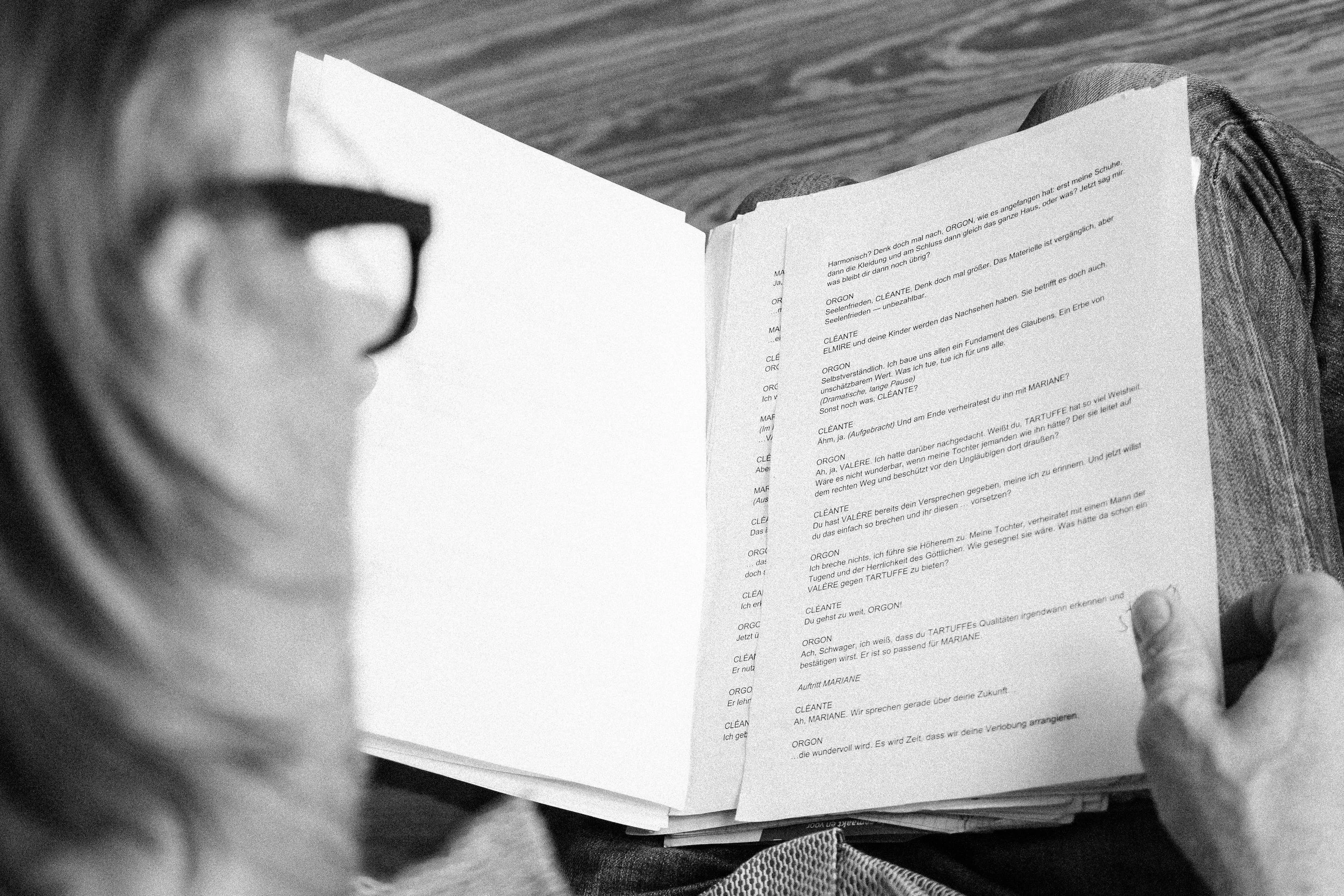 Eine Person liest ein Skript oder Drehbuch in schwarz-weiß, gehalten in der Hand, mit Textzeilen und Charakternamen auf der Seite, während sie sitzt, auf einem Holztisch.