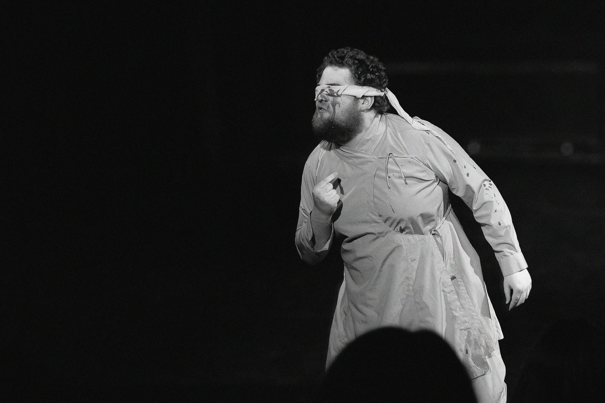 Schwarzes und weißes Bild eines Mannes in einer weißen Jacke, der mit verbundenen Augen und einem Lächeln steht, vermutlich bei einer Performance oder Inszenierung.