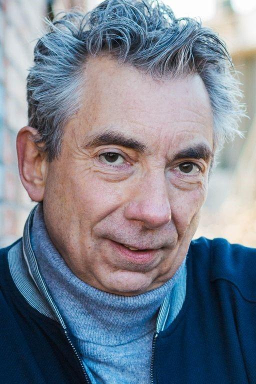Nahaufnahme eines älteren Mannes mit grauen, windgepeitschten Haaren, grauen Augen und einem leichten Lächeln, trägt einen blauen Pullover mit Reißverschluss und einen grauen Rollkragen darunter, im Freien bei Tageslicht.