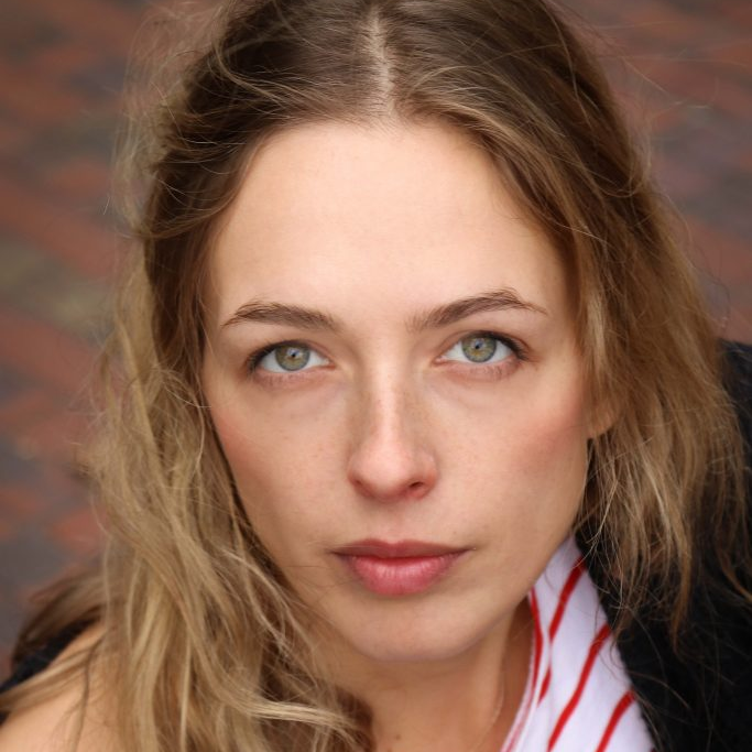 Nahaufnahme eines jungen Mädchens mit blonden, gewellten Haaren und blauen Augen, blickt direkt in die Kamera, im Hintergrund ein gepflasterter Boden.
