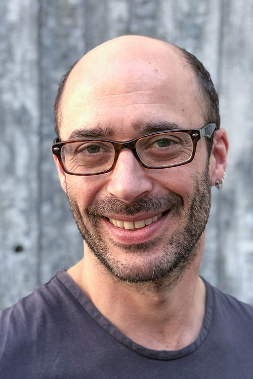 Ein Mann mit Brille, kurzen dunklen Haaren und einem Lächeln fotografiert vor einer Holzgestaltung im Hintergrund.