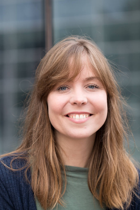 Janika Görres - Dozentin der internationalen Schauspiel-Akademie für Film und Theater in Hamburg. Junge Frau mit braunen Haaren und blauen Augen lächelt vor einem modernen Gebäude.