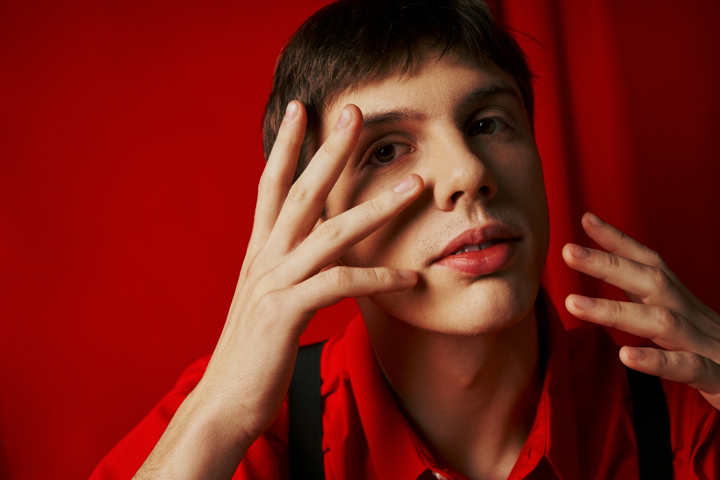 Nahaufnahme eines jungen Mannes mit kurzen Haaren vor rotem Hintergrund, der die Hand vor sein Gesicht hält.