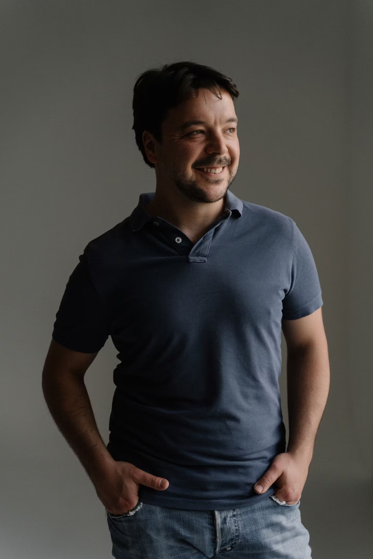 Ein lächelnder Mann trägt ein dunkelblaues Poloshirt und Jeans gegen einen neutralen grauen Hintergrund.