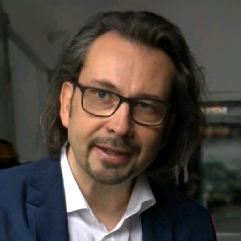 Lars Ceglecki - Dozent der internationalen Schauspiel-Akademie für Film und Theater in Hamburg. Mann mit langen Haaren, Brille und Krawatte in einem Raum, die Person spricht oder was sagt.