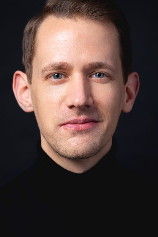 Johannes P. Braun - Dozent der internationalen Schauspiel-Akademie in Hamburg. Porträt eines jungen Mannes mit blonden Haaren, blauen Augen und einem schwarzen Rollkragenpullover vor dunklem Hintergrund.