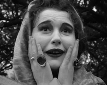 Schwarz-Weiß-Foto einer Frau mit verwunderten Ausdruck, Händen an Gesicht, große Ringe, dunkle Lippen, umgeben von Bäumen.