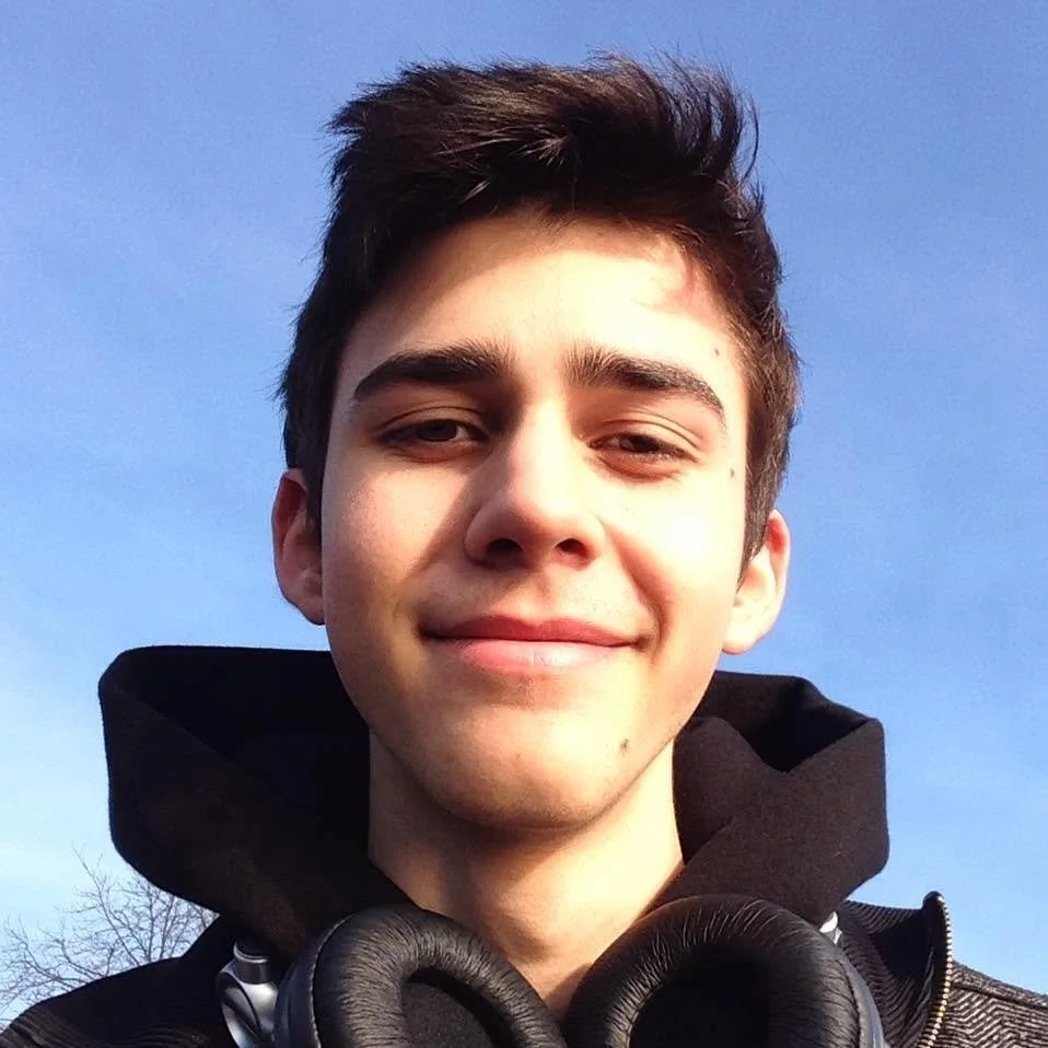 Ein junger Mann mit dunklem Haar trägt eine schwarze Jacke und Kopfhörer, steht vor einem blauen Himmel.