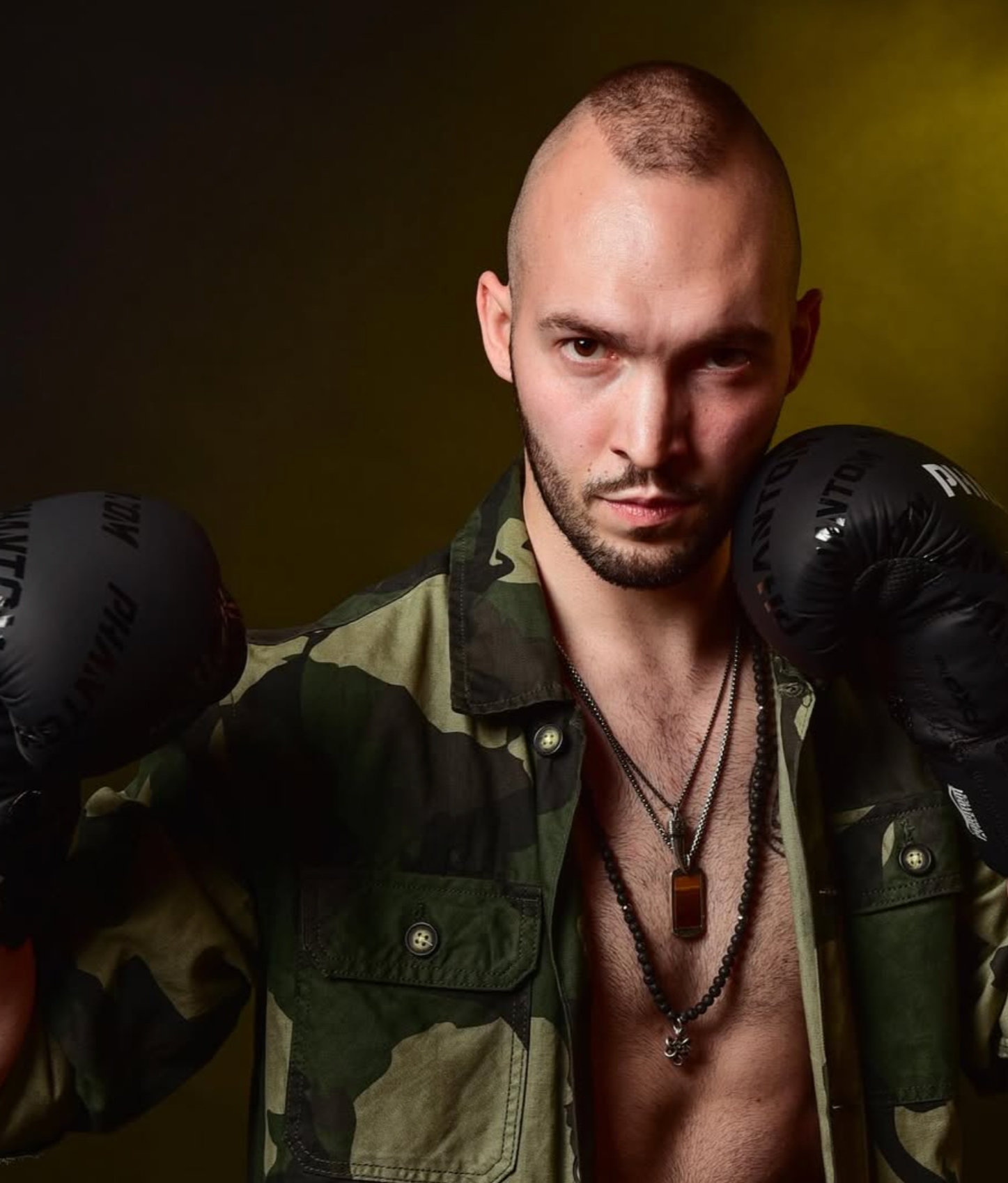 Luca Schenk - Dozent der internationalen Schauspiel-Akademie für Film und Theater in Hamburg. Ein Mann mit kahlköpfigem Haar, Bart, schwarze Boxhandschuhe, Camouflage-Jacke, offene Hemd, mehrere Halsketten, vor grün-gelbem Hintergrund.