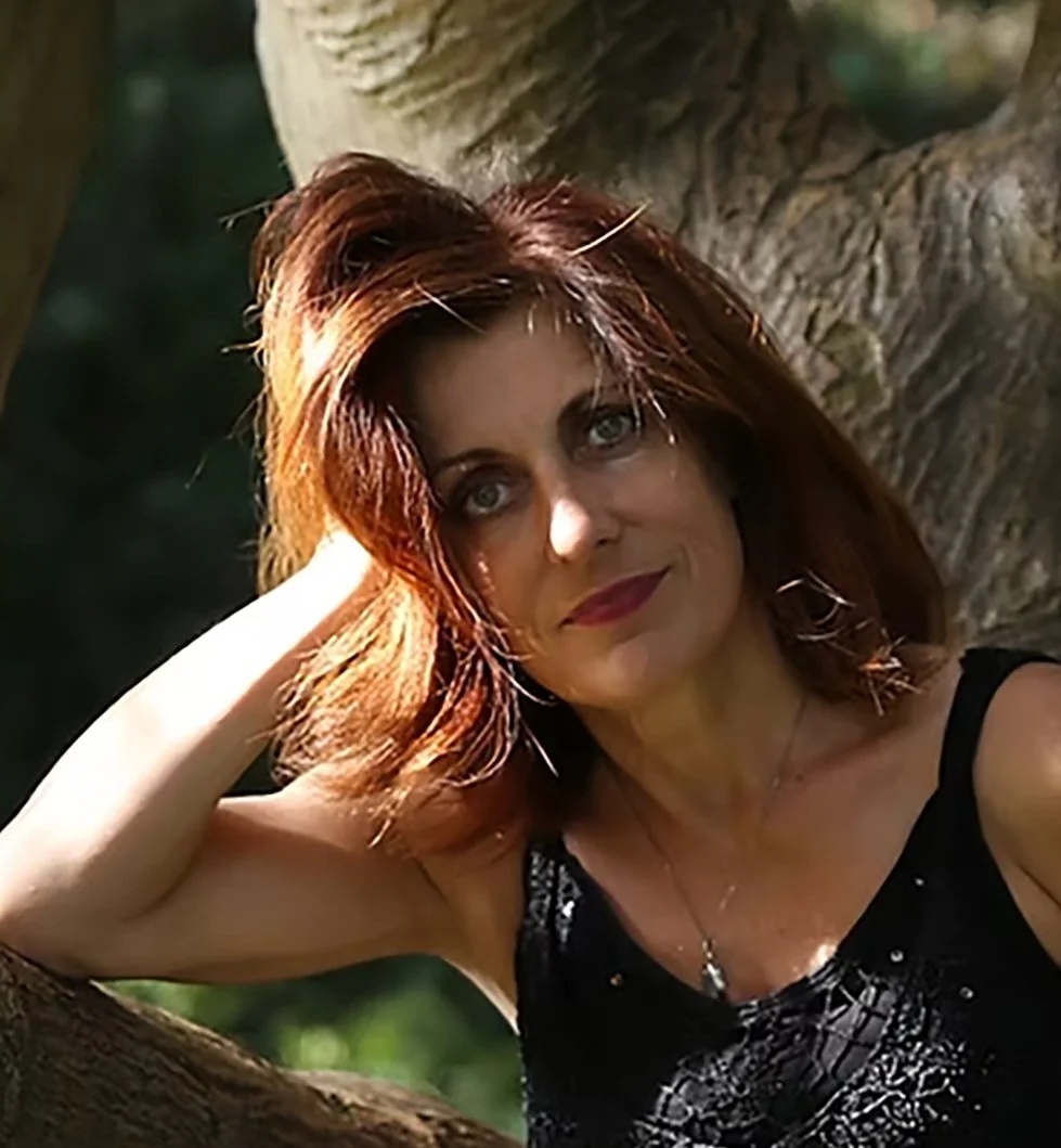 Eine Frau mit roten Haaren sitzt entspannt an einem Baum im Freien, schaut in die Kamera und lächelt leicht.