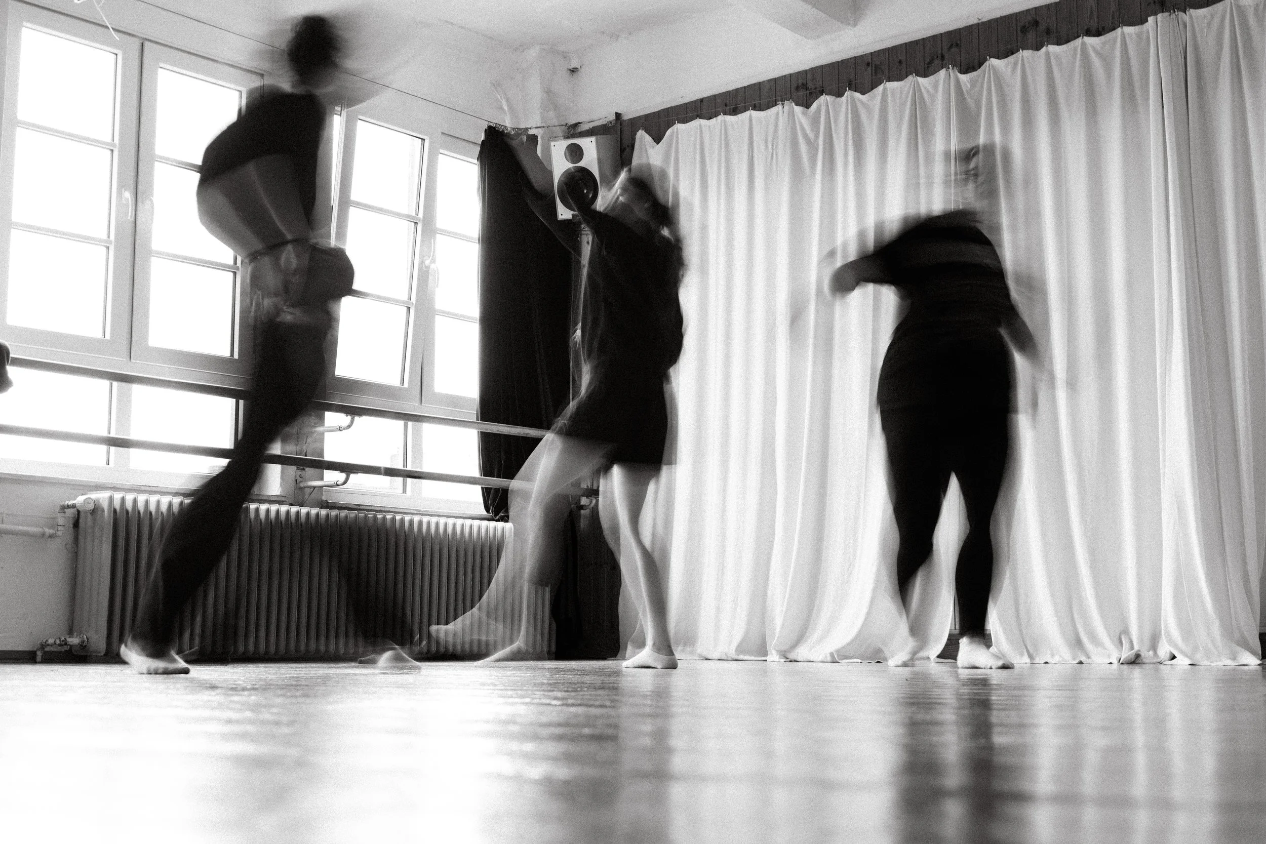 Balletttänzer in Bewegung im Tanzstudio, schwarz-weiß Foto, große Fenster, Vorhang im Hintergrund, Holzfußboden.