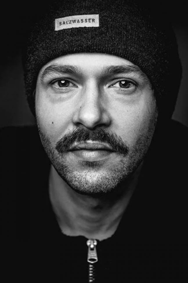Friedtjof Bundel - Dozent der internationalen Schauspiel-Akademie für Film und Theater in Hamburg. Schwarz-weiß-Fotografie eines Mannes mit Bart, der eine schwarze Mütze trägt mit Aufnäher 'Salzwasser'