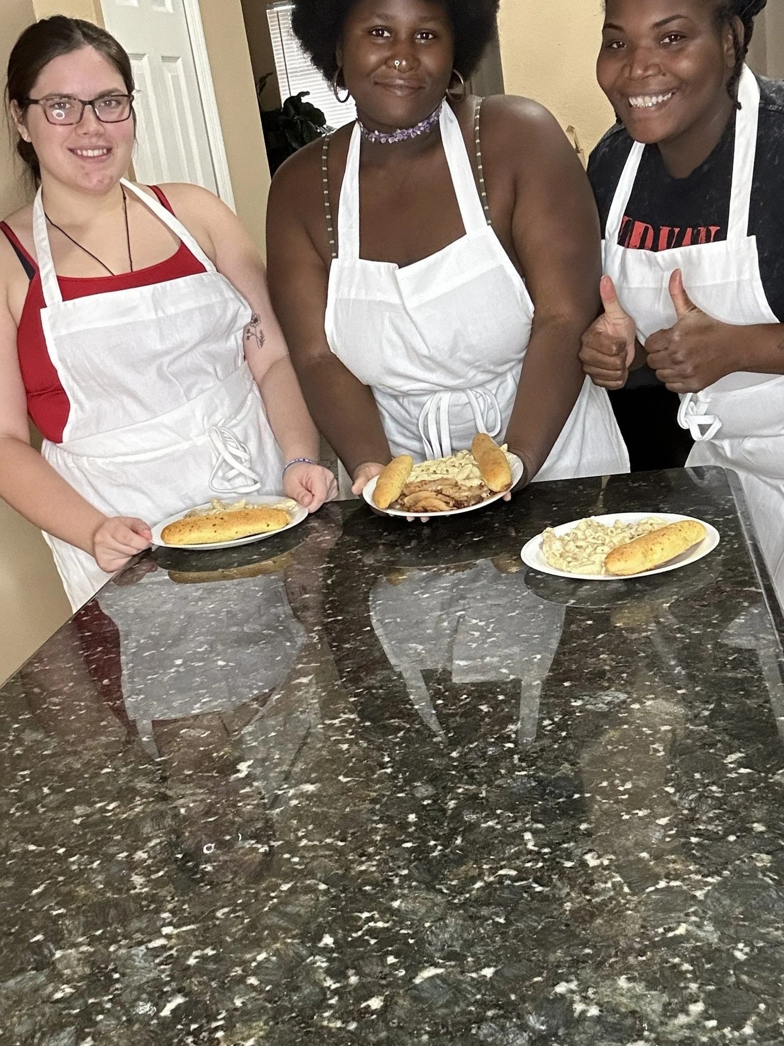 Three young women wearing white aprons standing behind a kitchen counter with plates of food, smiling and giving thumbs up.