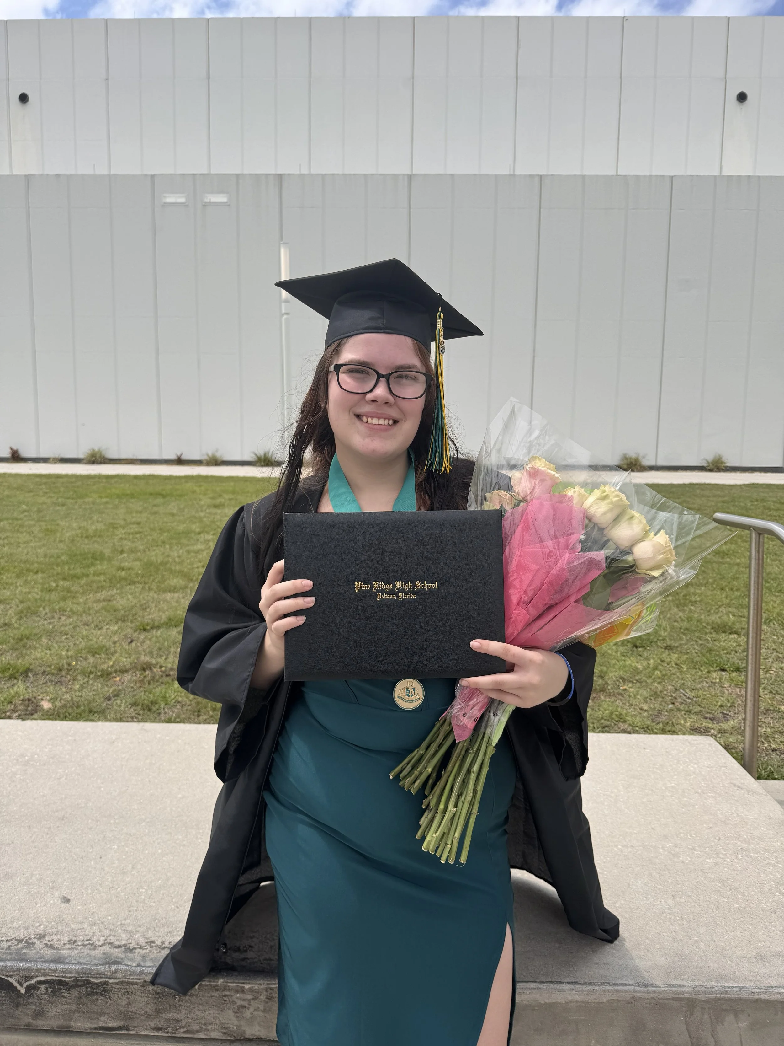 Young woman in a teal dress and black graduation gown holding a bouquet of pink and white roses and a diploma cover outdoors.